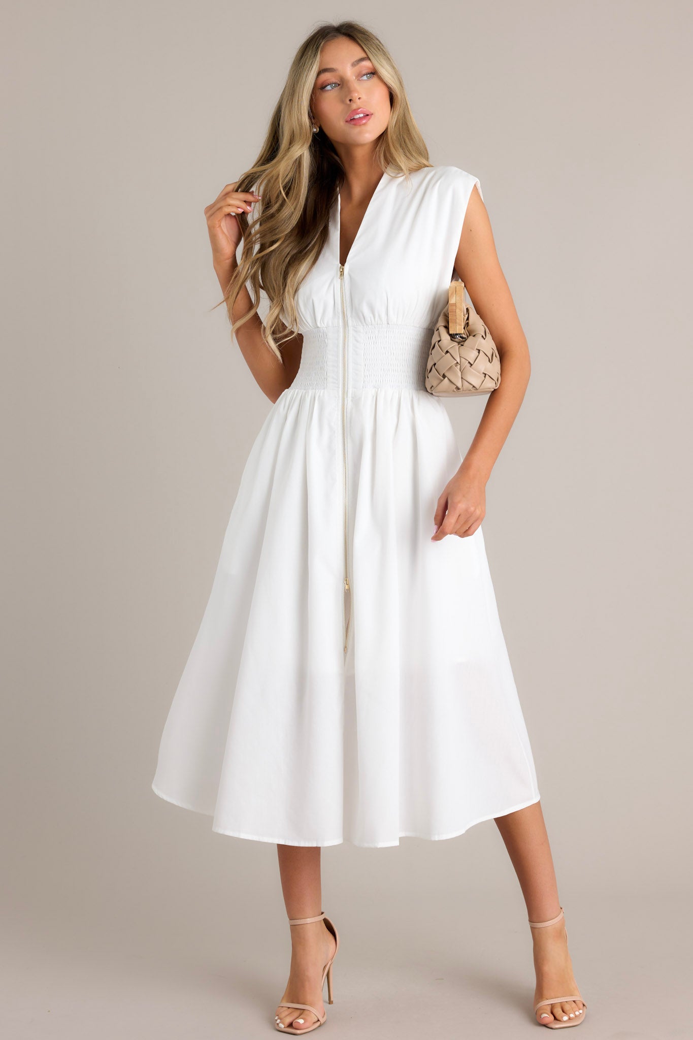Front view of a white dress with a V-neckline, cap sleeves, a zippered front, and a cinched waist with shirred detailing, featuring a flowing midi-length skirt with a front slit, highlighting its elegant and stylish design.