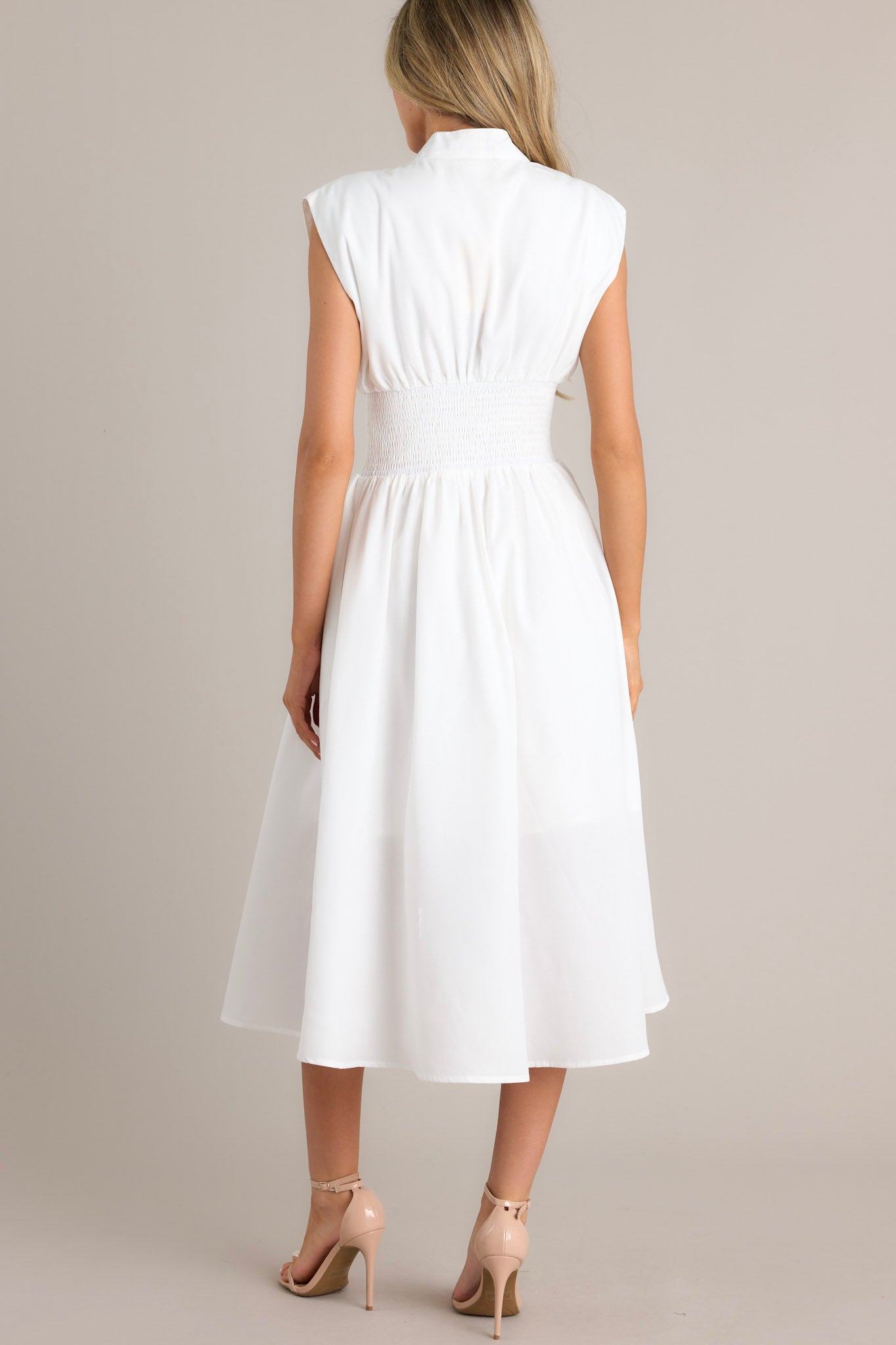 Back view of this white dress with a V-neckline, cap sleeves, a zippered front, and a cinched waist with shirred detailing, featuring a flowing midi-length skirt with a front slit.