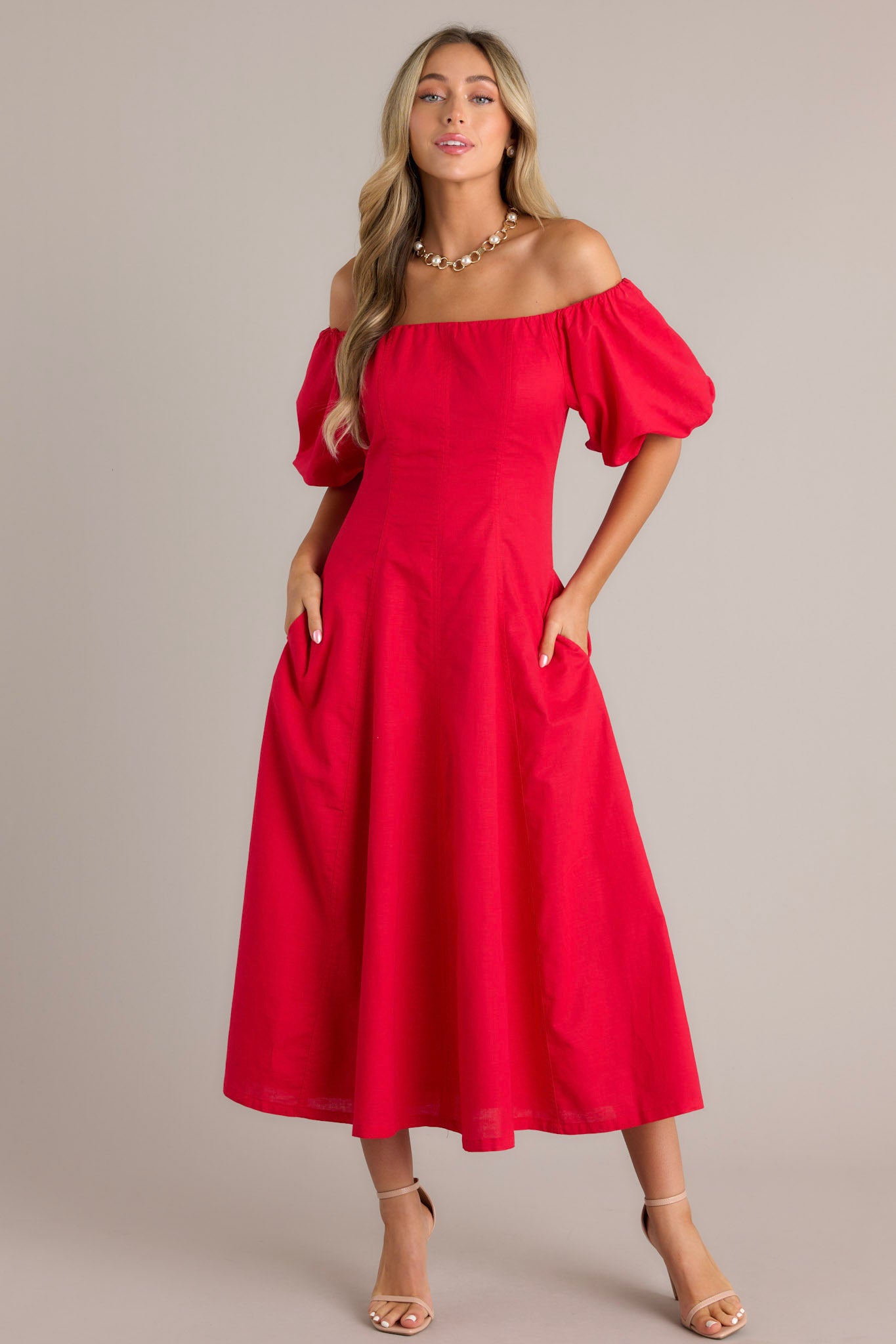 Full body view of this off-the-shoulder red dress featuring puffed sleeves and a fitted bodice that flares out into a flowy, ankle-length skirt.