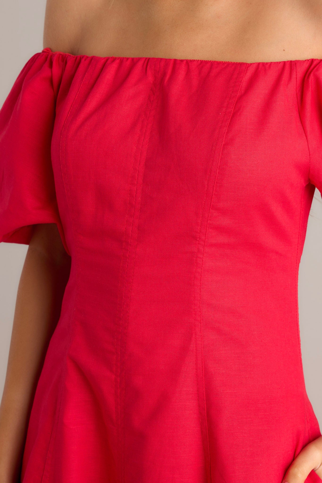 Close up view of this red midi dress that features an off shoulder elastic neckline, a discrete back zipper, functional hip pockets, visible front and back seams, elastic cuffed puff half sleeves, and a flowing silhouette.