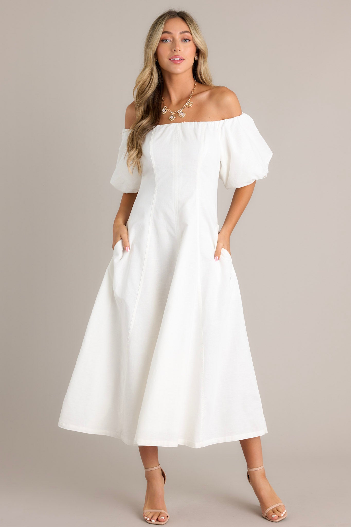 Front view of an elegant off-the-shoulder white dress with voluminous puffed sleeves, a fitted bodice, and a flowing ankle-length skirt, featuring side pockets for added functionality.