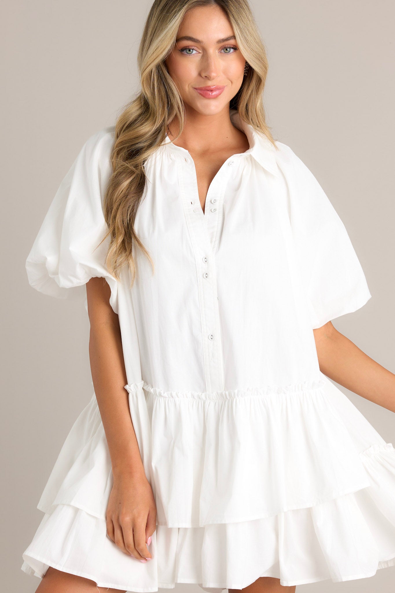 Close up front view of this White mini dress with a collared neckline, button-up front, and short puff sleeves. The dress has a tiered, ruffled skirt, giving it a playful and chic style.