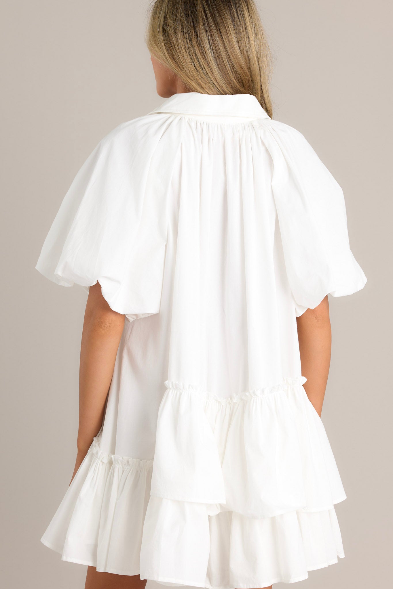 Back view of this White mini dress with a collared neckline, button-up front, and short puff sleeves. The dress has a tiered, ruffled skirt, giving it a playful and chic style.