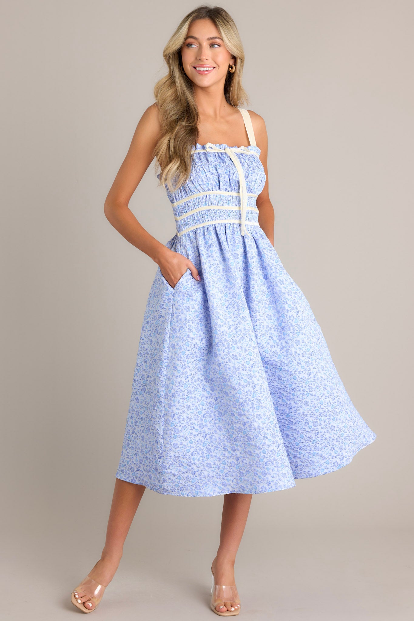 Full body view of this light blue midi dress that features a square neckline, thick adjustable straps, fully smocked bust and waist, a self-tie bust feature, velvet accents, functional hip pockets, and embossment throughout.