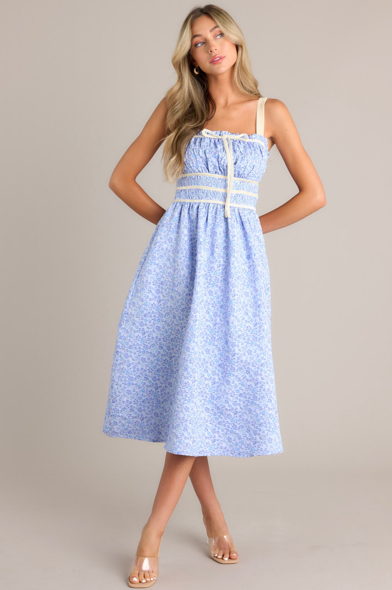 Front view of this light blue midi dress that features a square neckline, thick adjustable straps, fully smocked bust and waist, a self-tie bust feature, velvet accents, functional hip pockets, and embossment throughout.