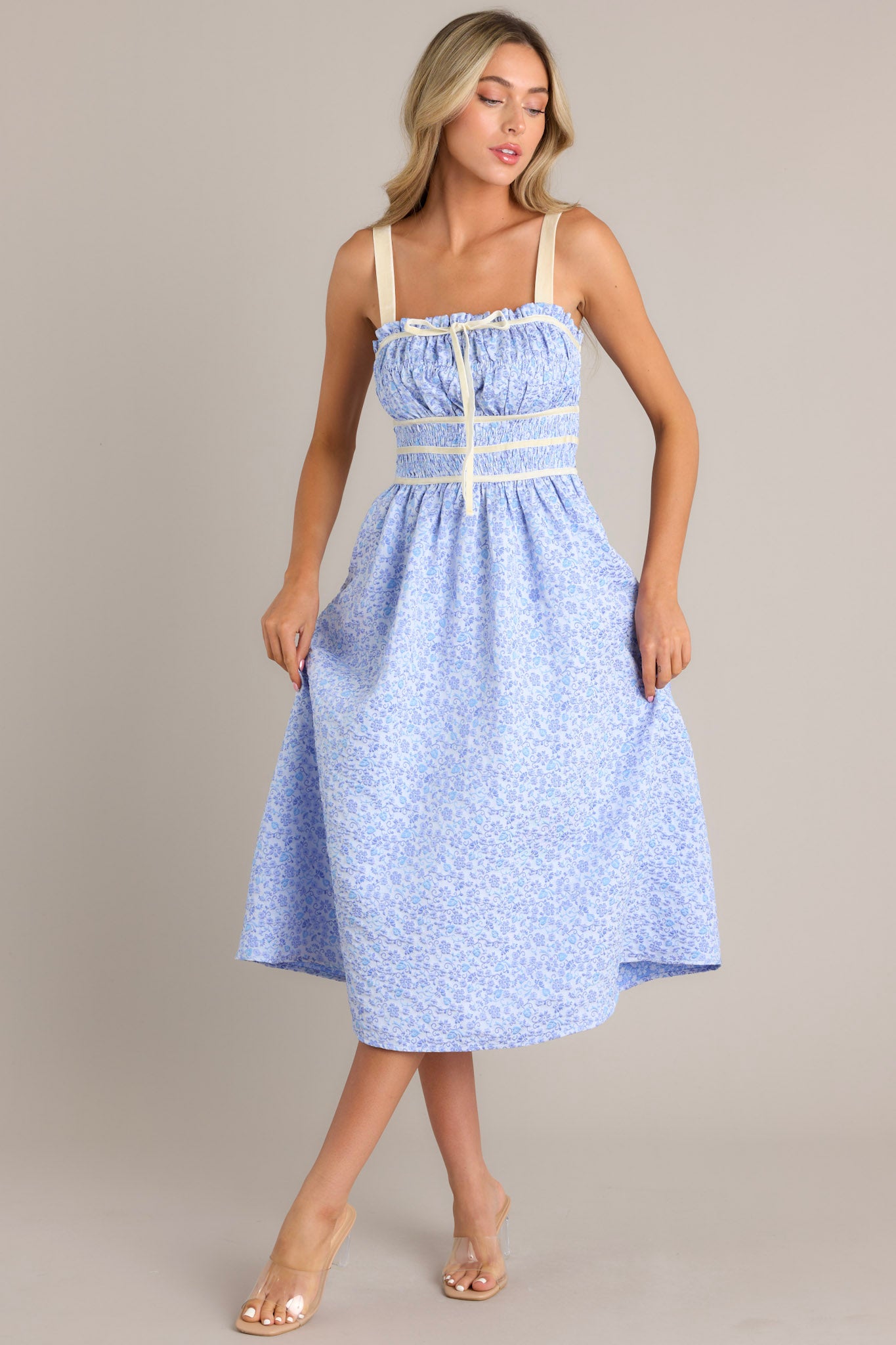 Angled front view of this light blue midi dress that features a square neckline, thick adjustable straps, fully smocked bust and waist, a self-tie bust feature, velvet accents, functional hip pockets, and embossment throughout.