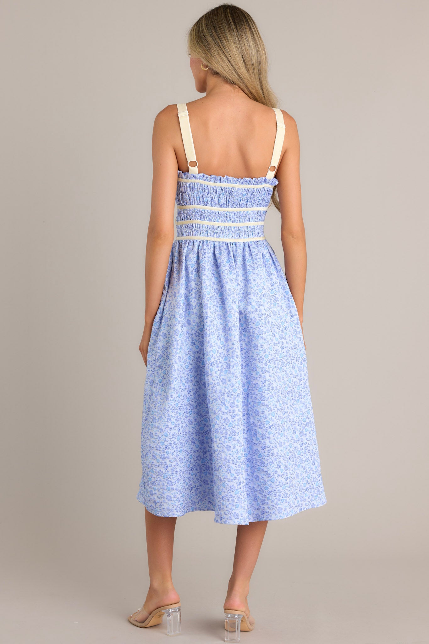 Back view of this light blue midi dress that features a square neckline, thick adjustable straps, fully smocked bust and waist, a self-tie bust feature, velvet accents, functional hip pockets, and embossment throughout.