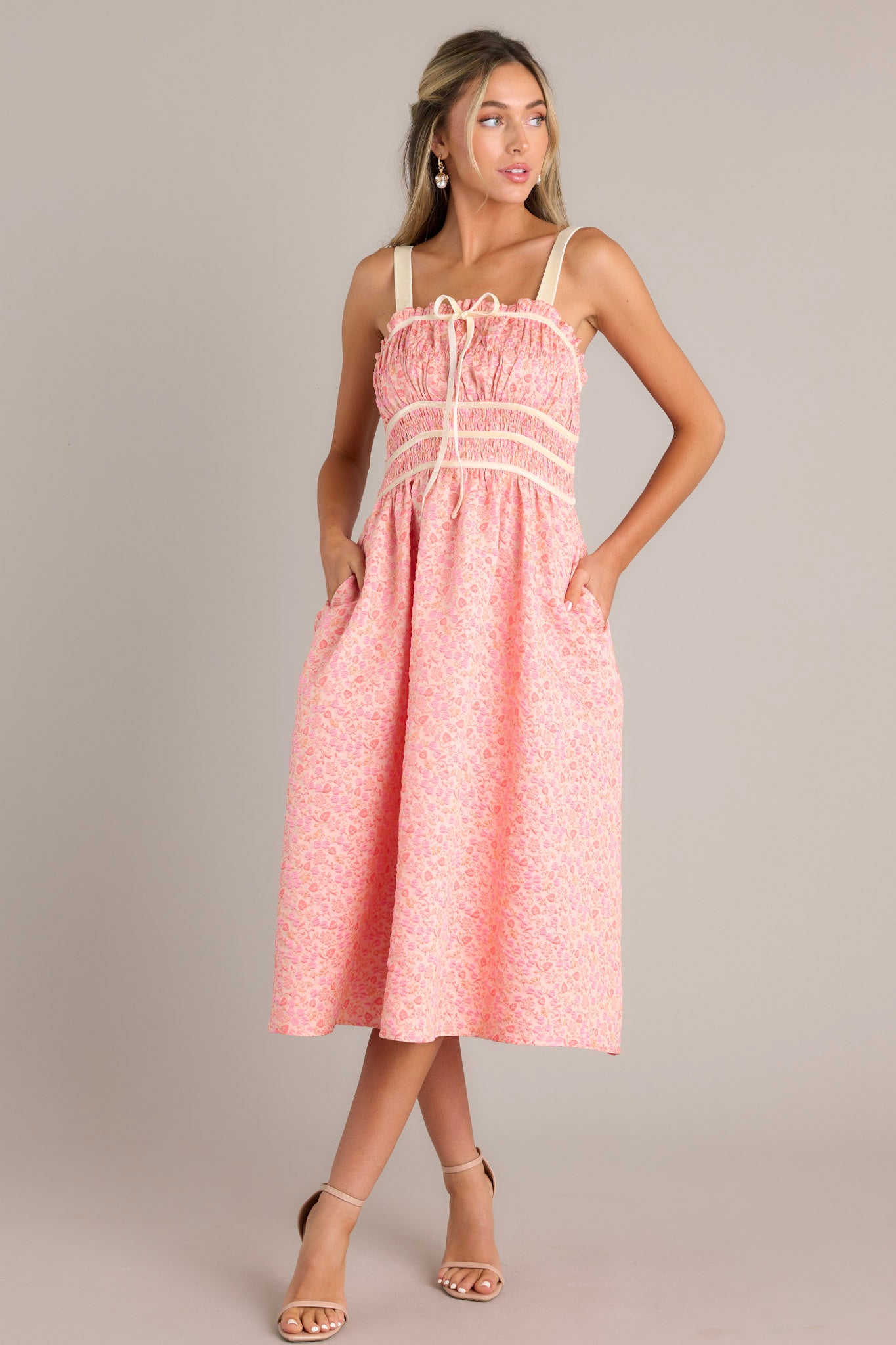 Front view of this light pink midi dress that features a square neckline, thick adjustable straps, fully smocked bust and waist, a self-tie bust feature, velvet accents, functional hip pockets, and embossment throughout.