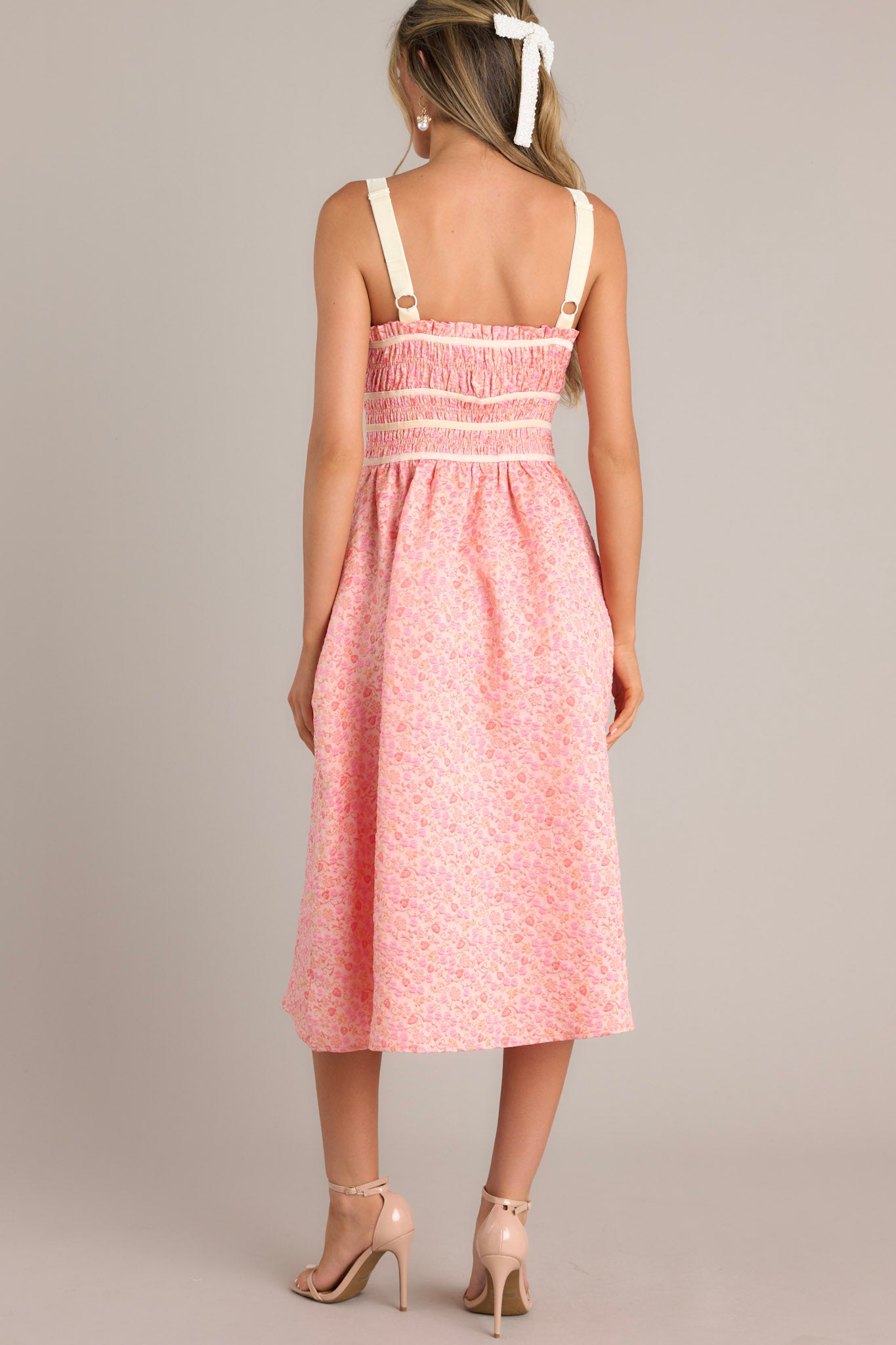 Back view of this light pink midi dress that features a square neckline, thick adjustable straps, fully smocked bust and waist, a self-tie bust feature, velvet accents, functional hip pockets, and embossment throughout.