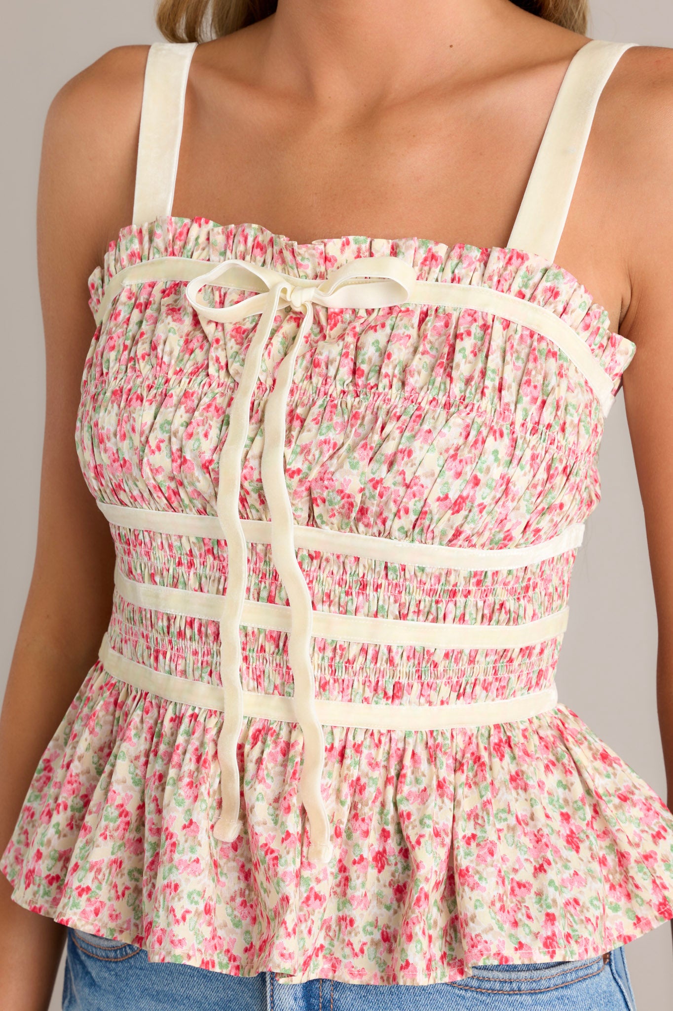 Detail shot of a sleeveless pink floral smocked top with a slight sweetheart neckline, a peplum hem, ribbon detailing along the waist and bust, and ribbon bow.