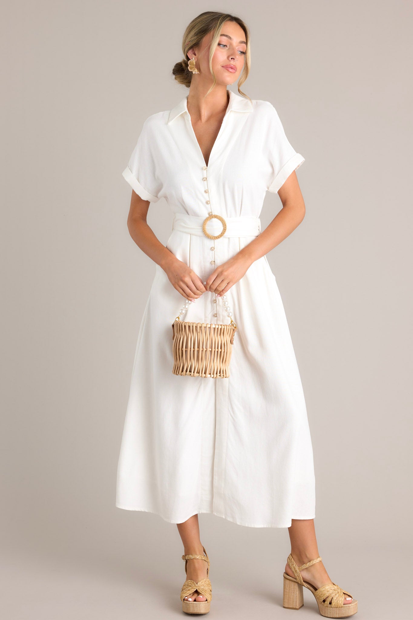 Angled full body view of this white maxi dress that features a collared v-neckline, a functional button front, a pull-through belt, functional hip pockets, a front slit, and cuffed short sleeves.