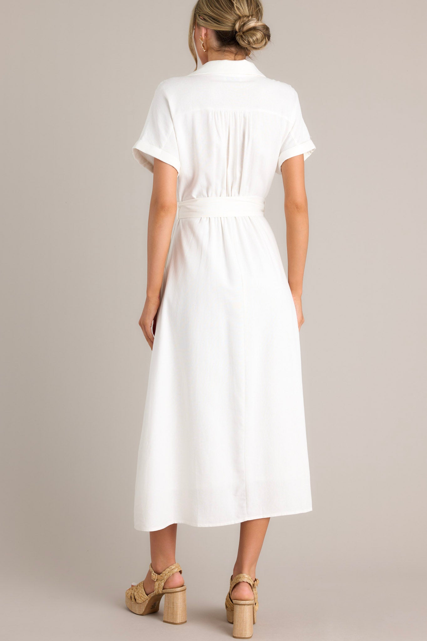 Back view of this white maxi dress that features a collared v-neckline, a functional button front, a pull-through belt, functional hip pockets, a front slit, and cuffed short sleeves.