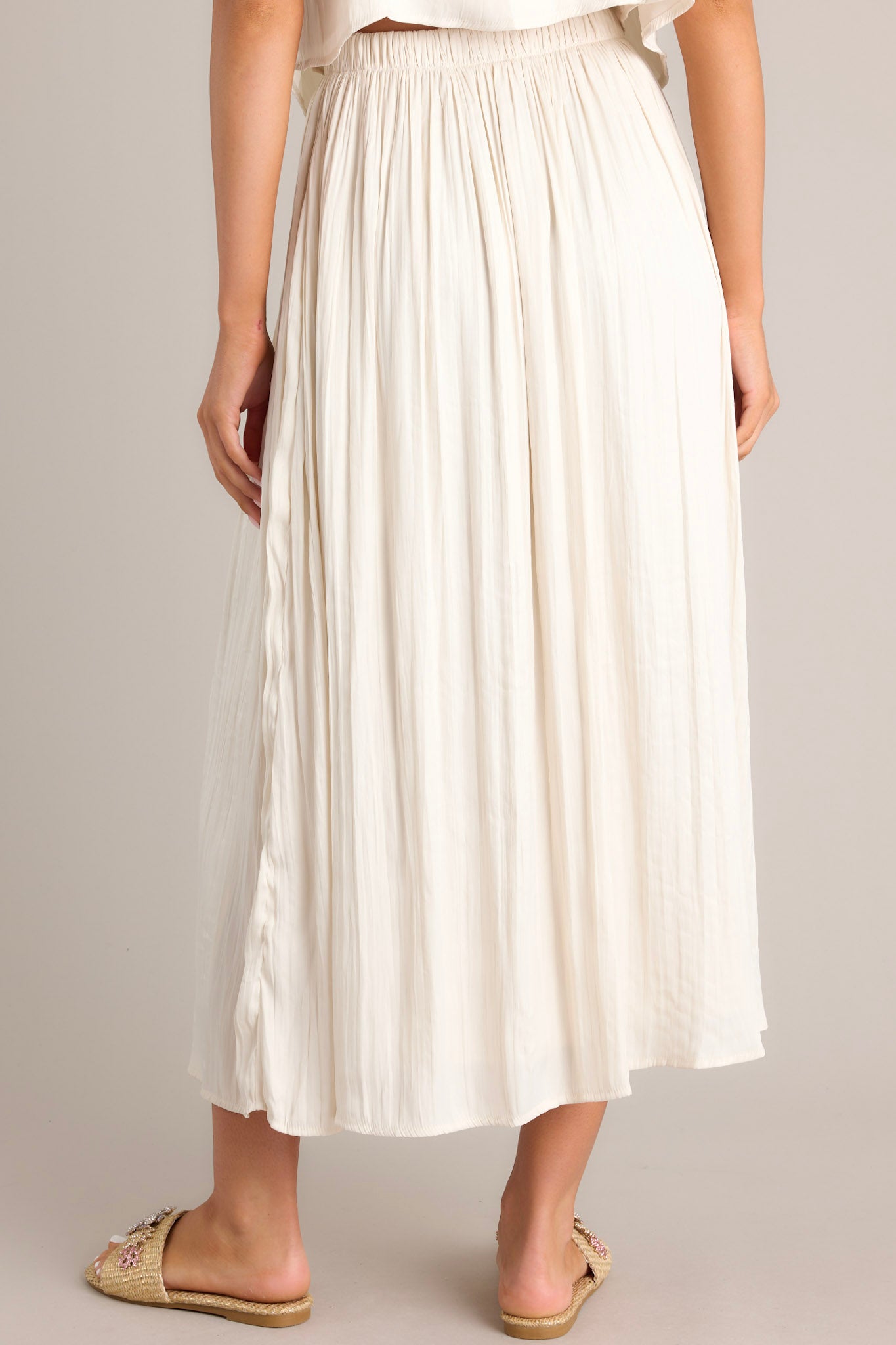 Back view of this midi sandstone skirt that features a high waisted design, an elastic waistband, subtle pleats throughout, and a flowing silhouette.