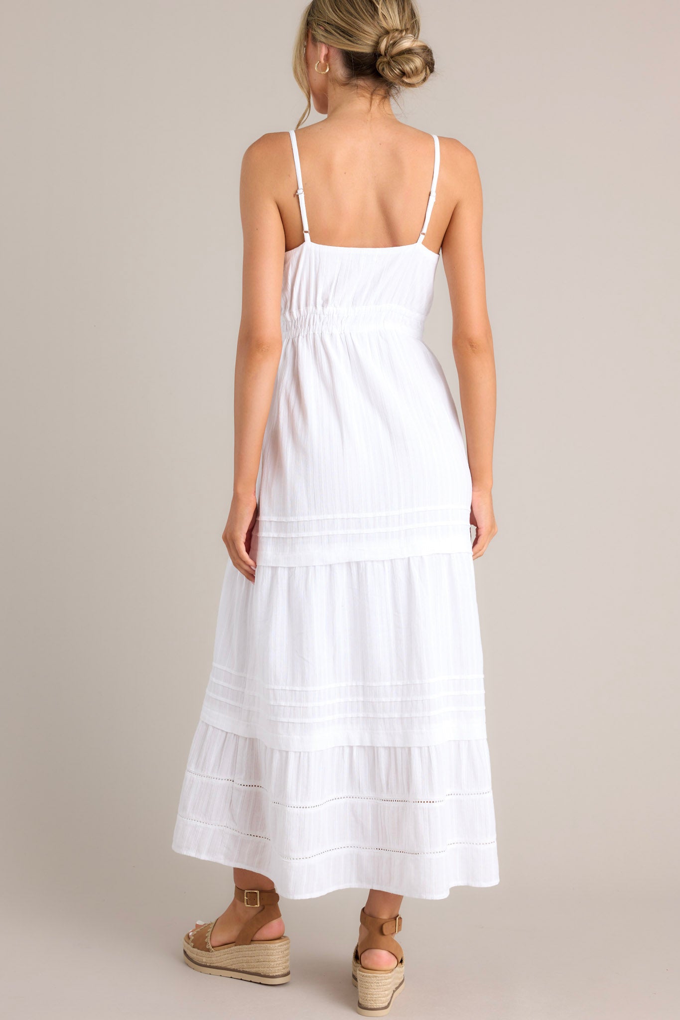 Back view of this white midi dress that features a v-neckline, thin adjustable straps, eyelet bust detailing, a self-tie bust feature, an elastic waistline, and a tiered design.