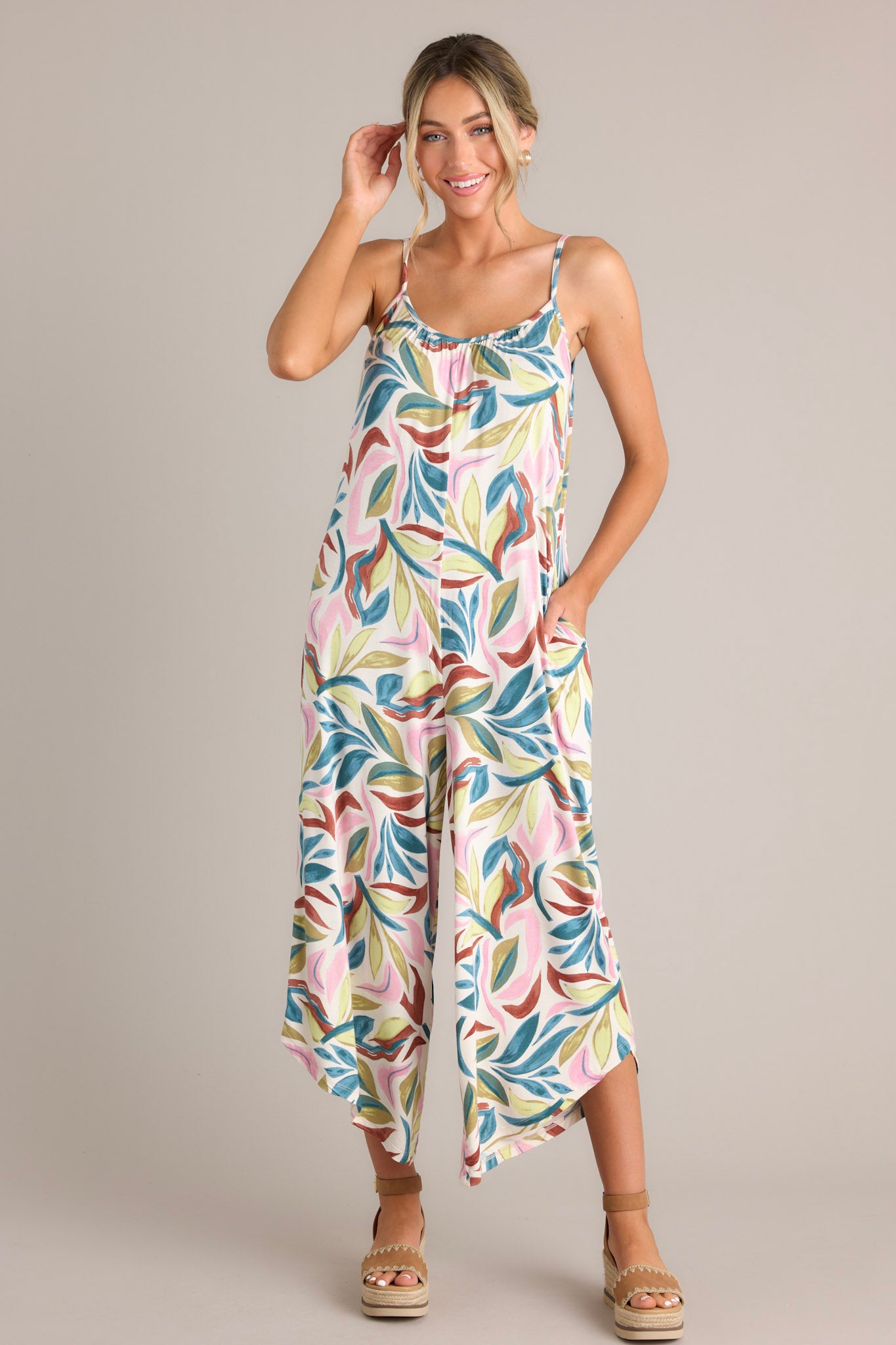 Action shot of a beige jumpsuit with a scoop neckline, thin adjustable straps, a relaxed fit, functional hip pockets, and a scooped hemline, highlighting the flow and movement of the jumpsuit.