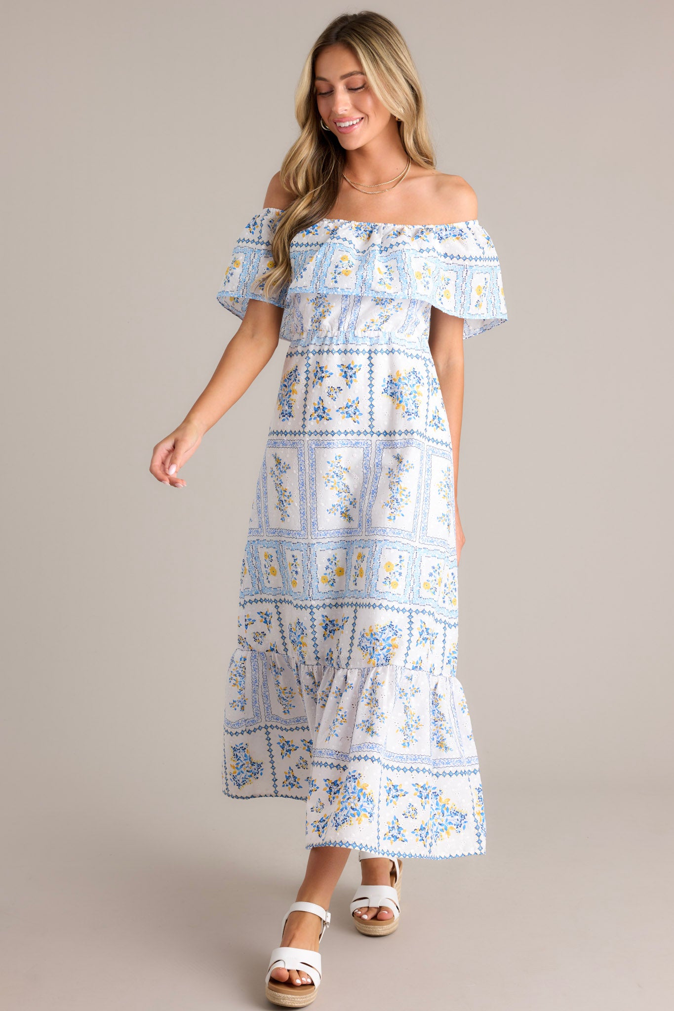 Front angled view of model walking in this white & blue maxi dress that features an elastic off-the-shoulder neckline, a frilled overlay that forms a false sleeve, an elastic stretch waist, an embroidered floral design, and a lined and tiered skirt.