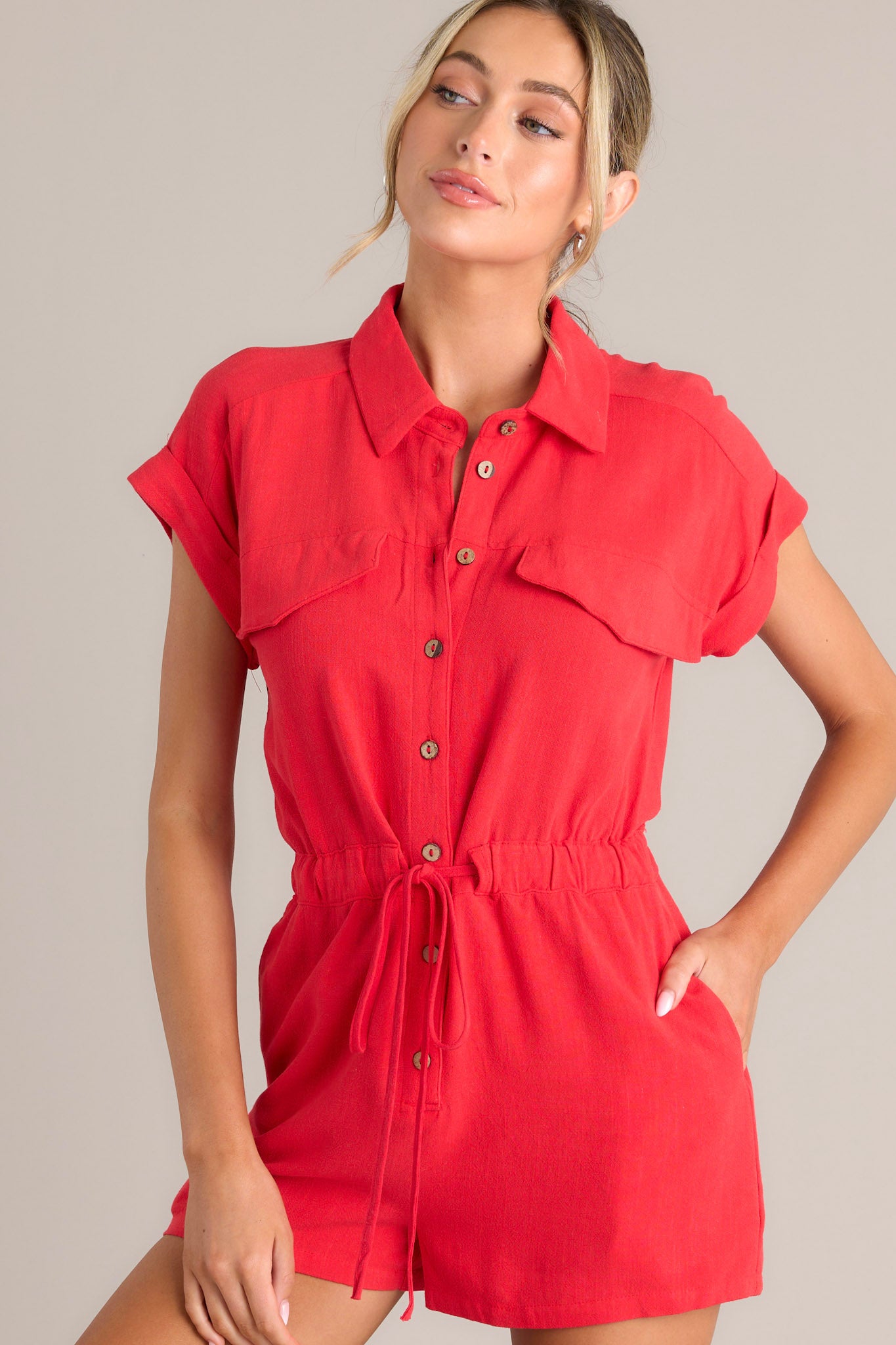 Angled front view of this red romper that features a collared neckline, a functional button front, faux breast pockets, an elastic waistband with a self-tie drawstring, functional hip pockets, and cuffed short sleeves.