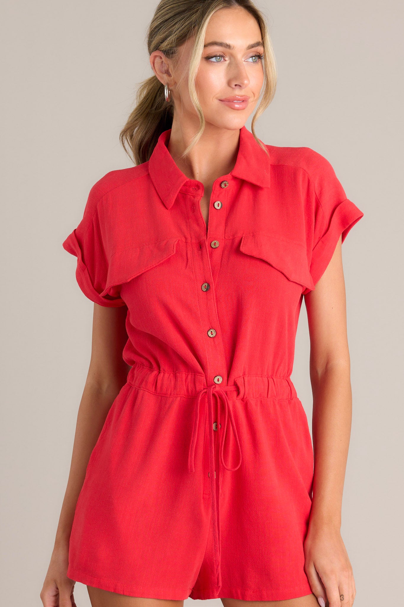 Front view of this red romper that features a collared neckline, a functional button front, faux breast pockets, an elastic waistband with a self-tie drawstring, functional hip pockets, and cuffed short sleeves.