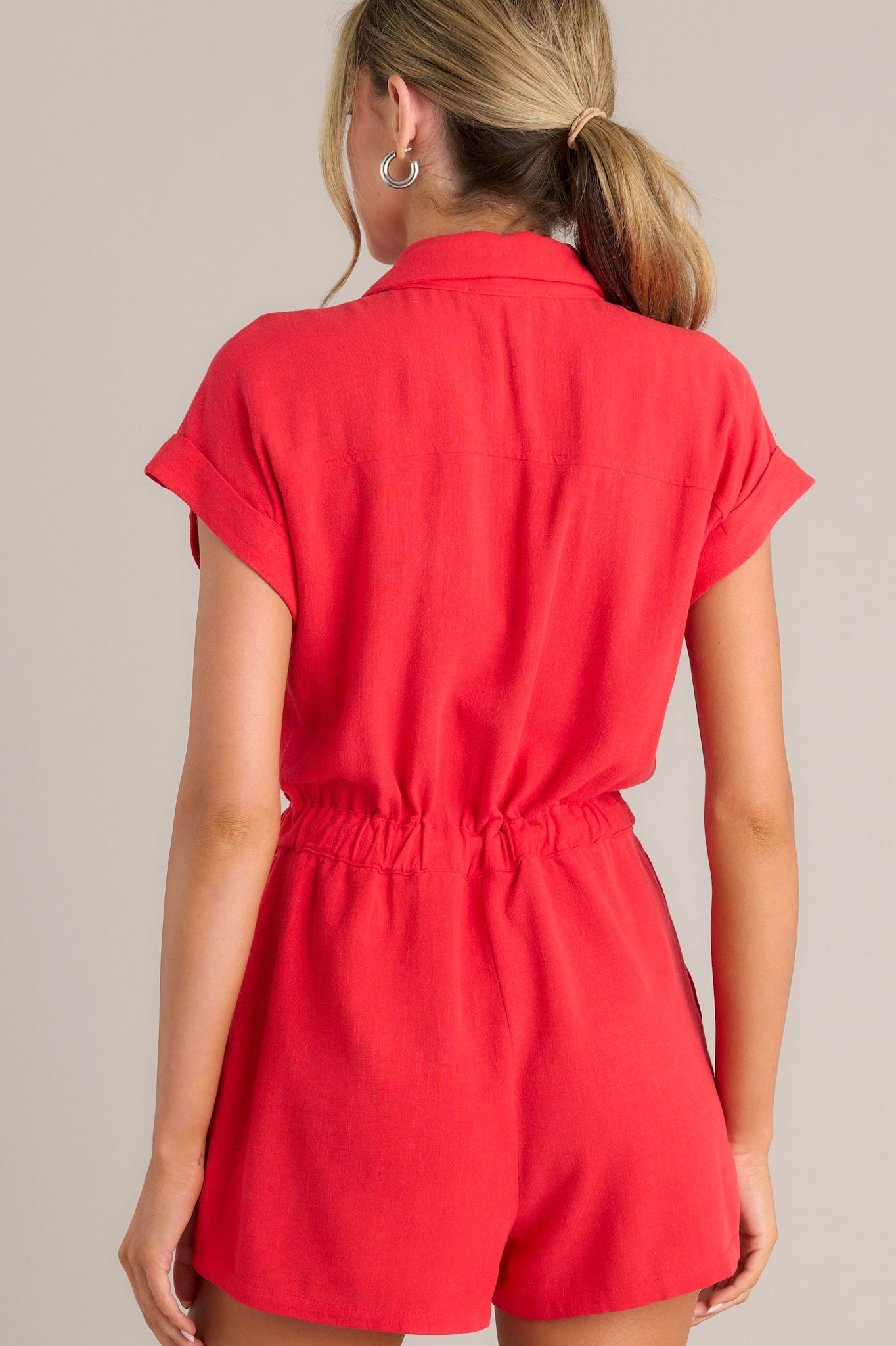 Back view of this red romper that features a collared neckline, a functional button front, faux breast pockets, an elastic waistband with a self-tie drawstring, functional hip pockets, and cuffed short sleeves.