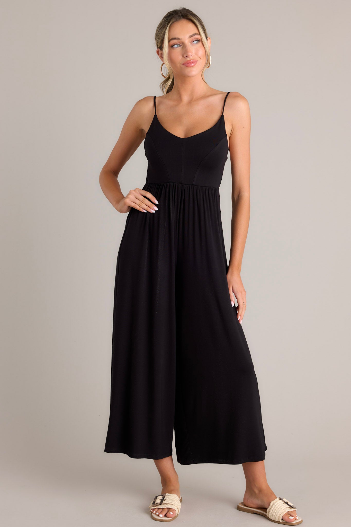 Front view of this black jumpsuit features a v-neckline, thin adjustable straps that cross in the back, an elastic waistband, and a wide leg.