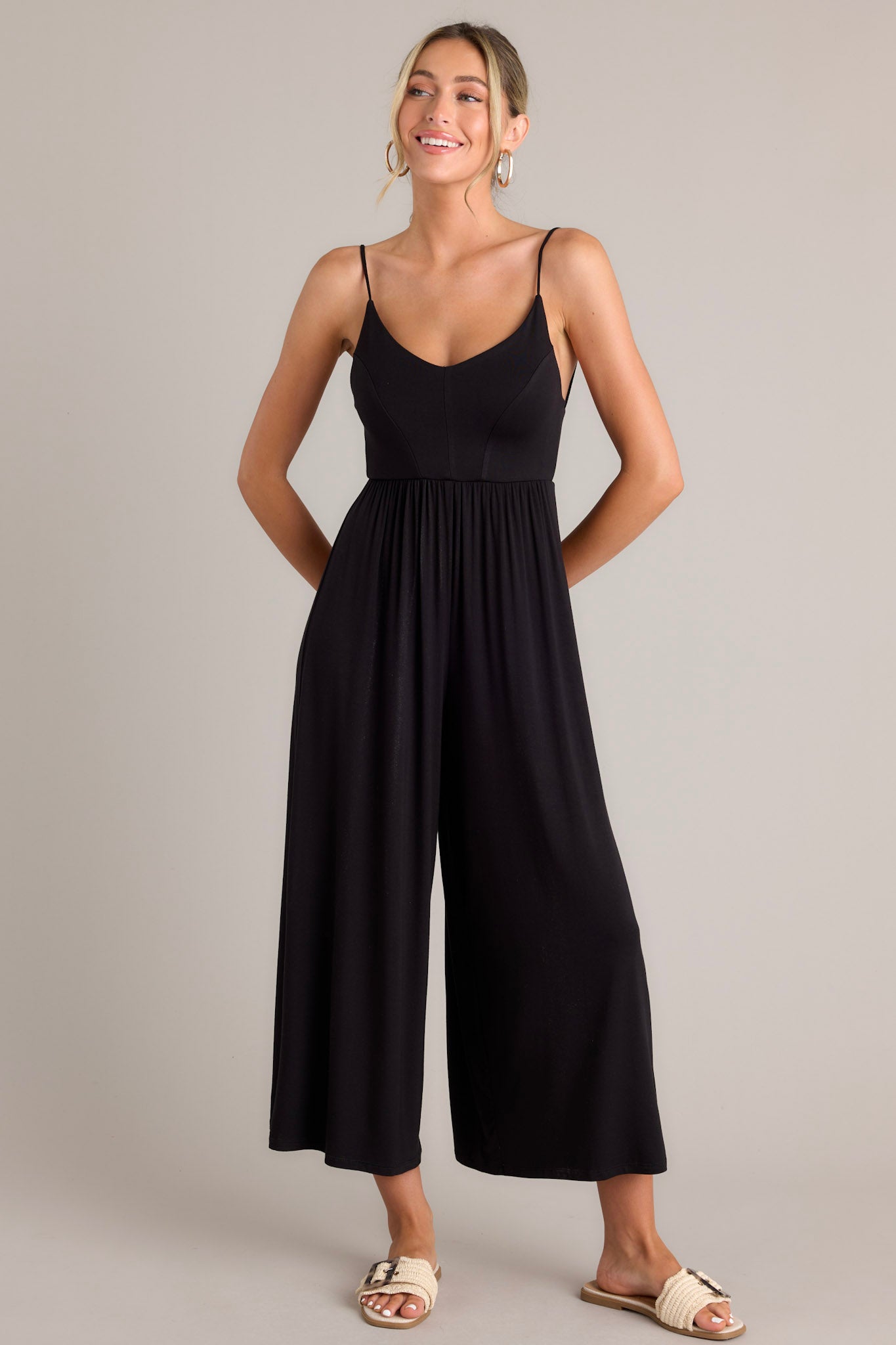 Full body view of this black jumpsuit that features a v-neckline, thin adjustable straps that cross in the back, an elastic waistband, and a wide leg.