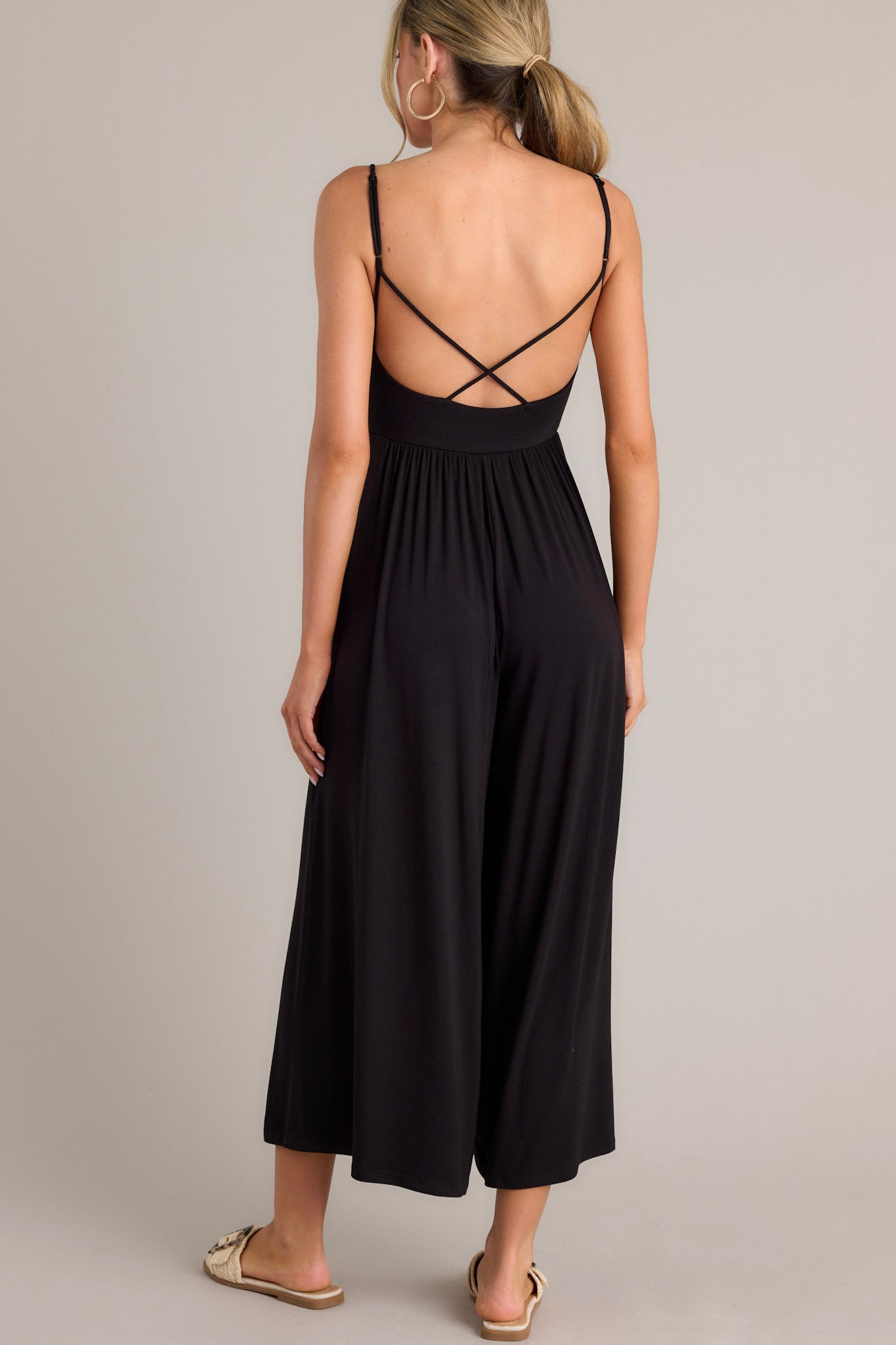 Back view of this black jumpsuit that features a v-neckline, thin adjustable straps that cross in the back, an elastic waistband, and a wide leg.