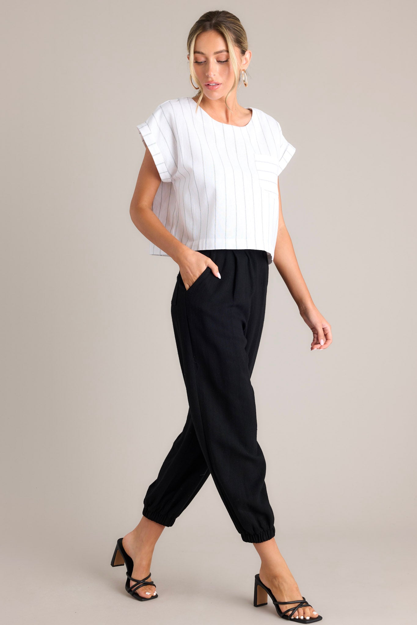 Side full body view of these black linen joggers that feature a high rise design, a functional button & zipper closure, belt loops, subtle pleats, functional front & back pockets, and elastic cuffed ankles