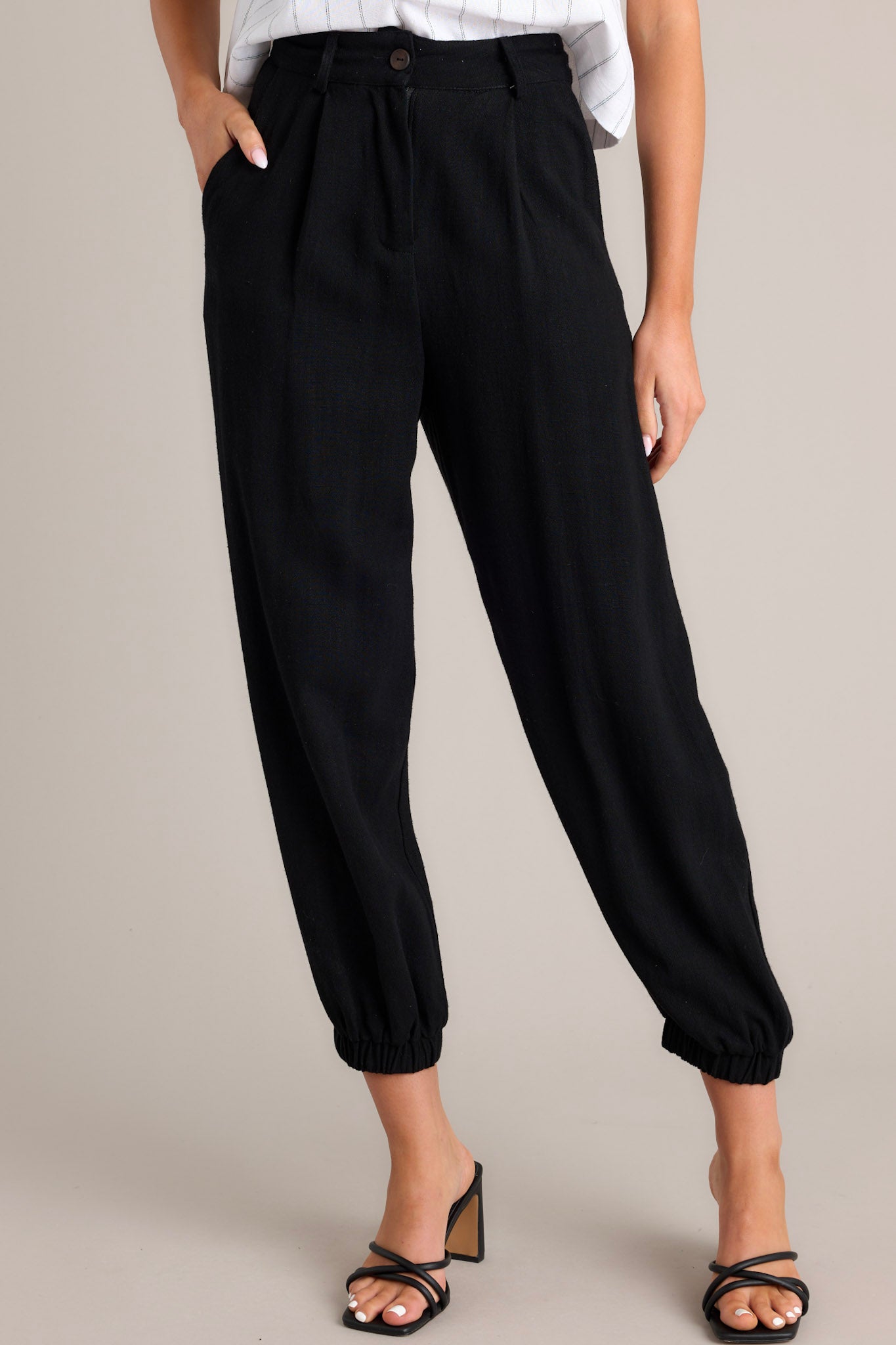 Front view of these black linen joggers that feature a high rise design, a functional button & zipper closure, belt loops, subtle pleats, functional front & back pockets, and elastic cuffed ankles