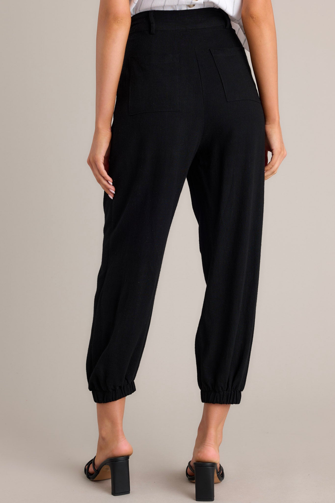 Back view of these black linen joggers that feature a high rise design, a functional button & zipper closure, belt loops, subtle pleats, functional front & back pockets, and elastic cuffed ankles