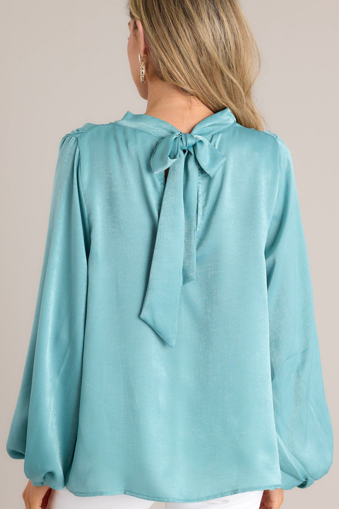 Back view of this seafoam blue top that features a high cowl neckline, a keyhole cutout at the back of the neck with a self-tie closure, balloon sleeves with buttoned cuffs, and a stunning shiny satin material. 