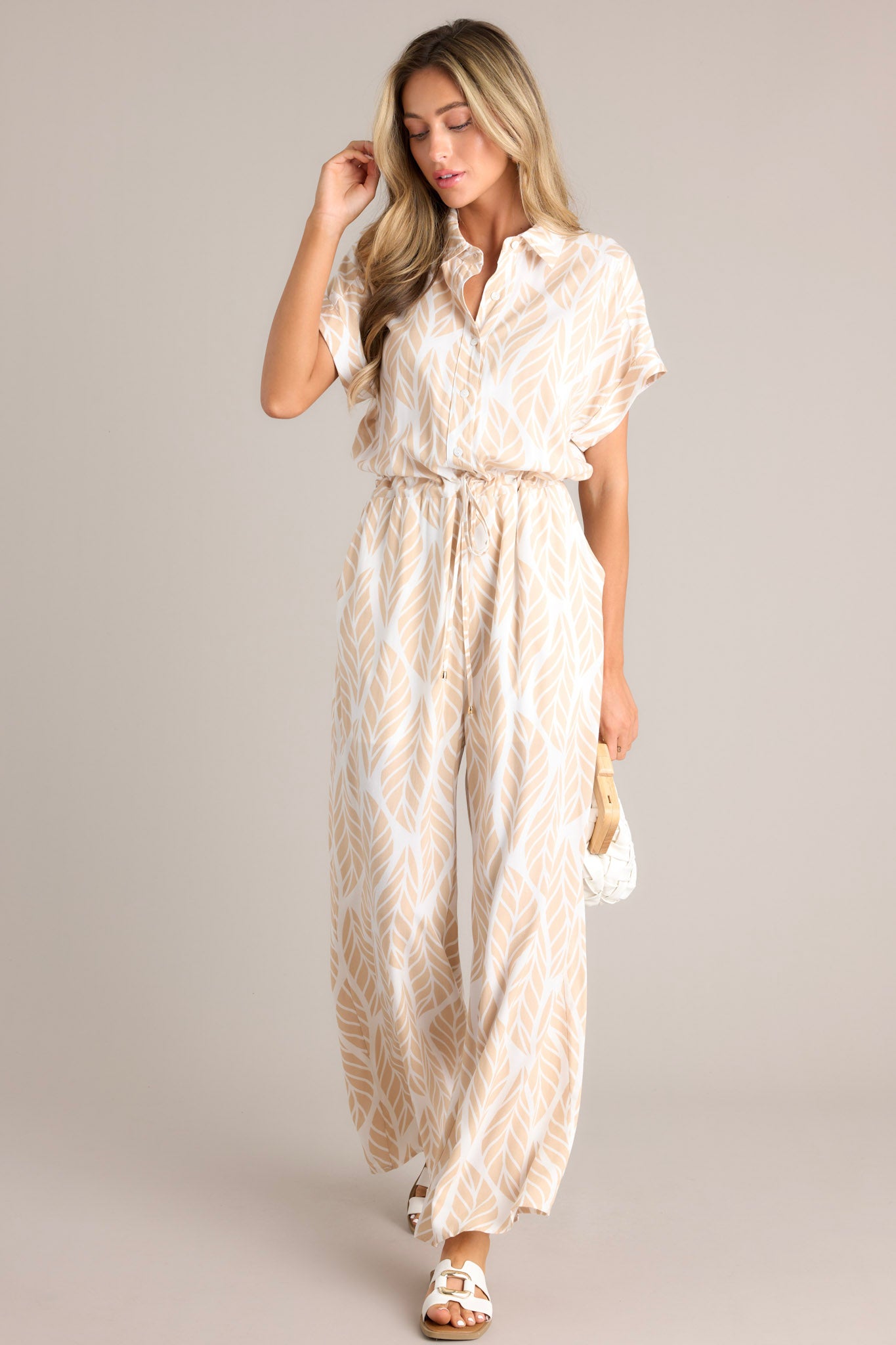 Angled full body view of this beige jumpsuit that features a collared neckline, cuffed short sleeves, a functional button front, a self-tie drawstring waist, functional hip pockets, and a wide leg.