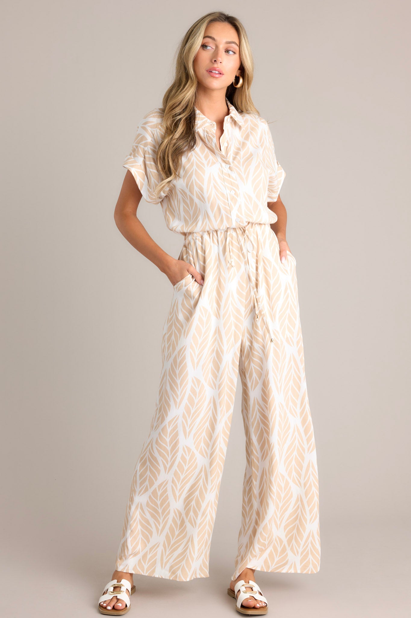 Angled front view of this beige jumpsuit that features a collared neckline, cuffed short sleeves, a functional button front, a self-tie drawstring waist, functional hip pockets, and a wide leg.