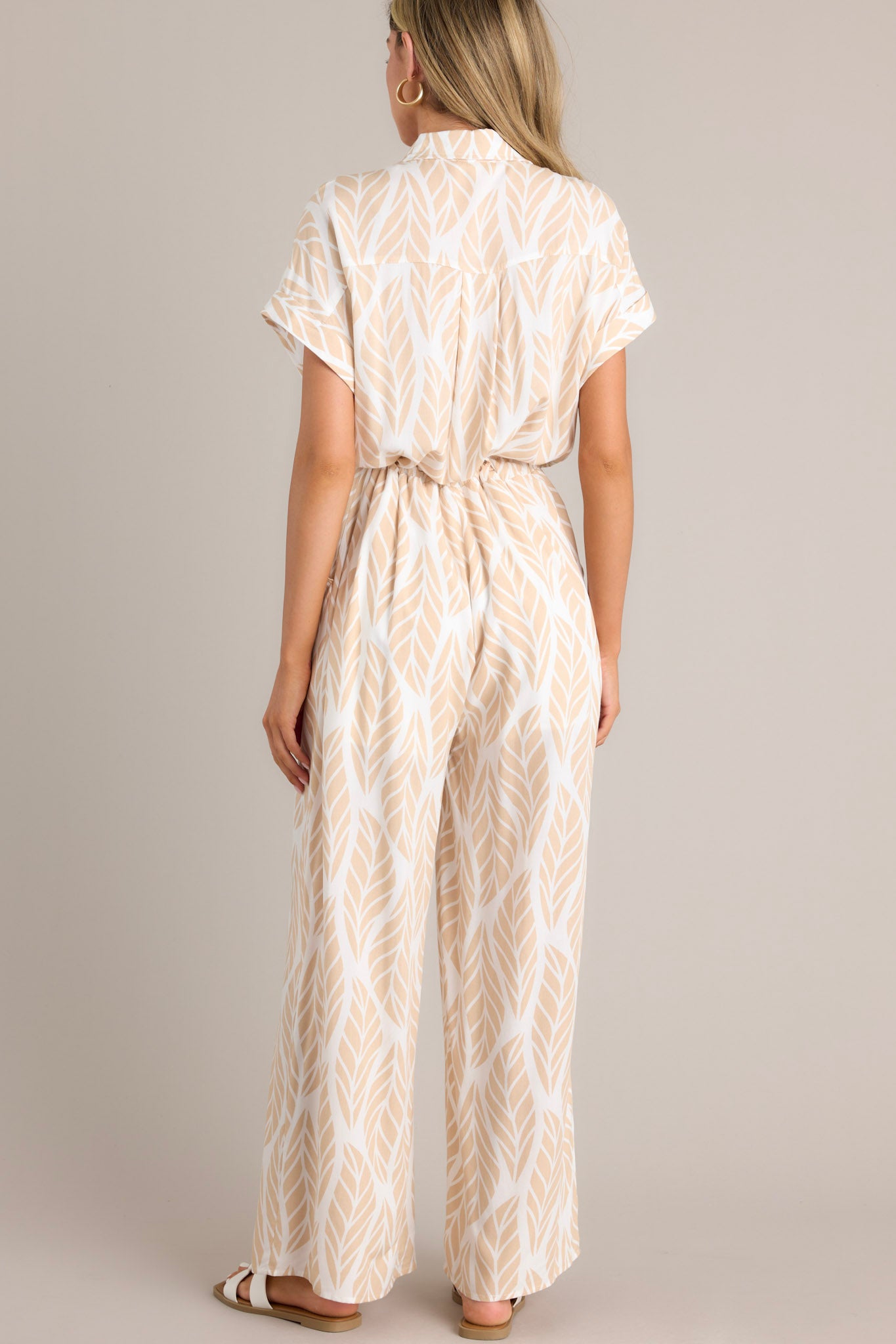 Back view of this beige jumpsuit that features a collared neckline, cuffed short sleeves, a functional button front, a self-tie drawstring waist, functional hip pockets, and a wide leg.