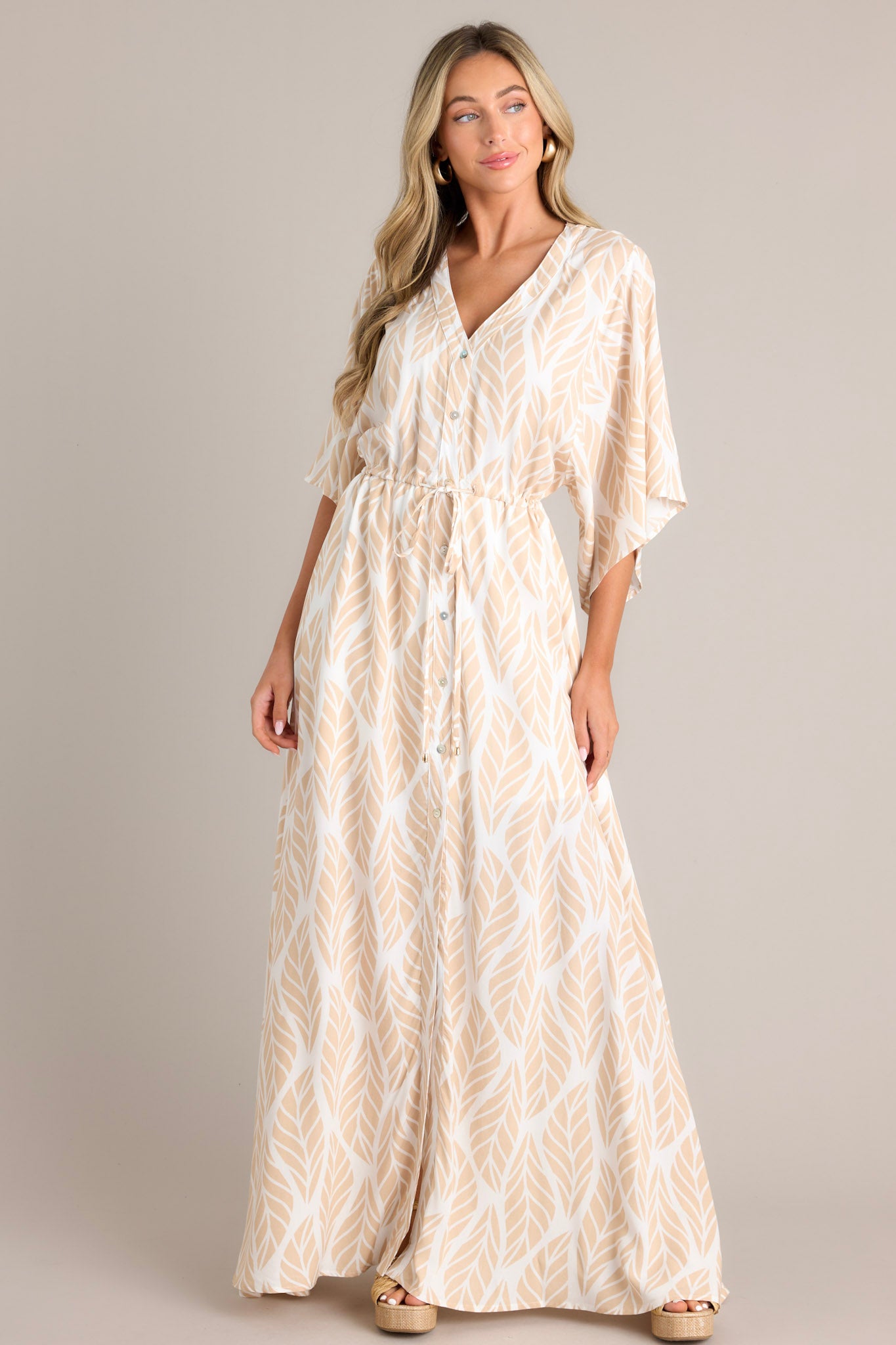 Full body front  view of this beige maxi dress that features a v-neckline, a functional button front, a self-tie drawstring waist, a flowing silhouette, and flowing half sleeves.