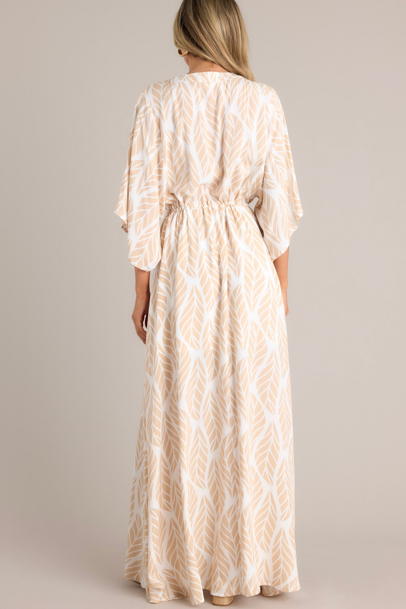 Back  view of this beige maxi dress that features a v-neckline, a functional button front, a self-tie drawstring waist, a flowing silhouette, and flowing half sleeves.