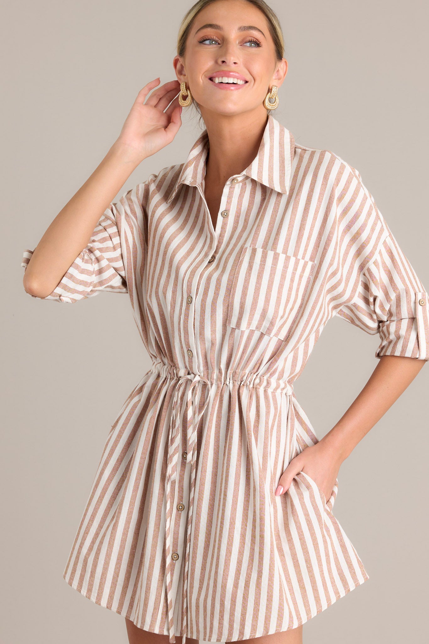 Front angled view of a stripe mini dress featuring a collared neckline, a full button front, a breast pocket, a self-tie drawstring waist, functional hip pockets, and button cuffed sleeves