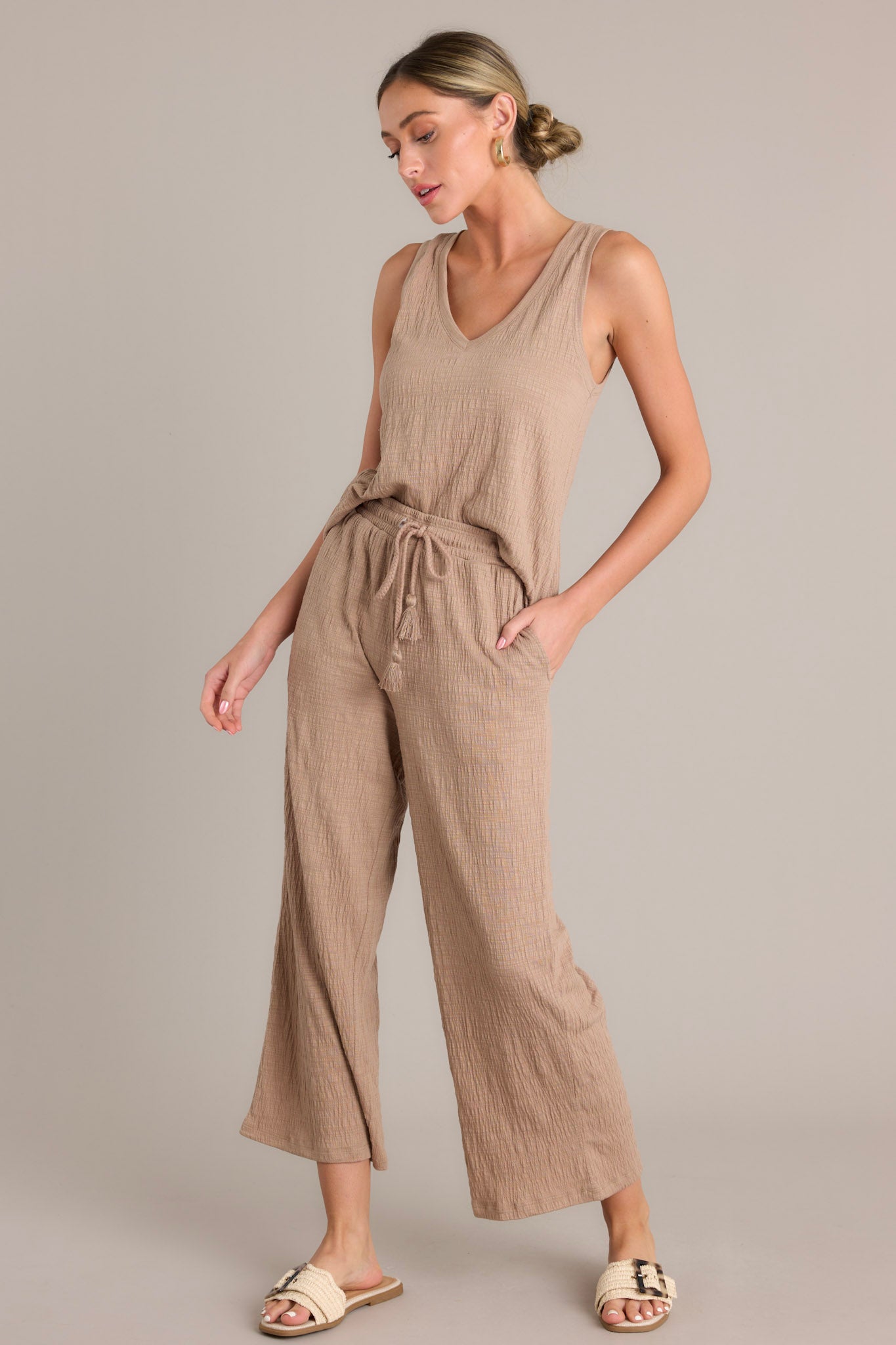 Front angled view of high-waisted beige pants featuring an elastic waistband, self-tie drawstring, functional hip pockets, textured material, and wide legs