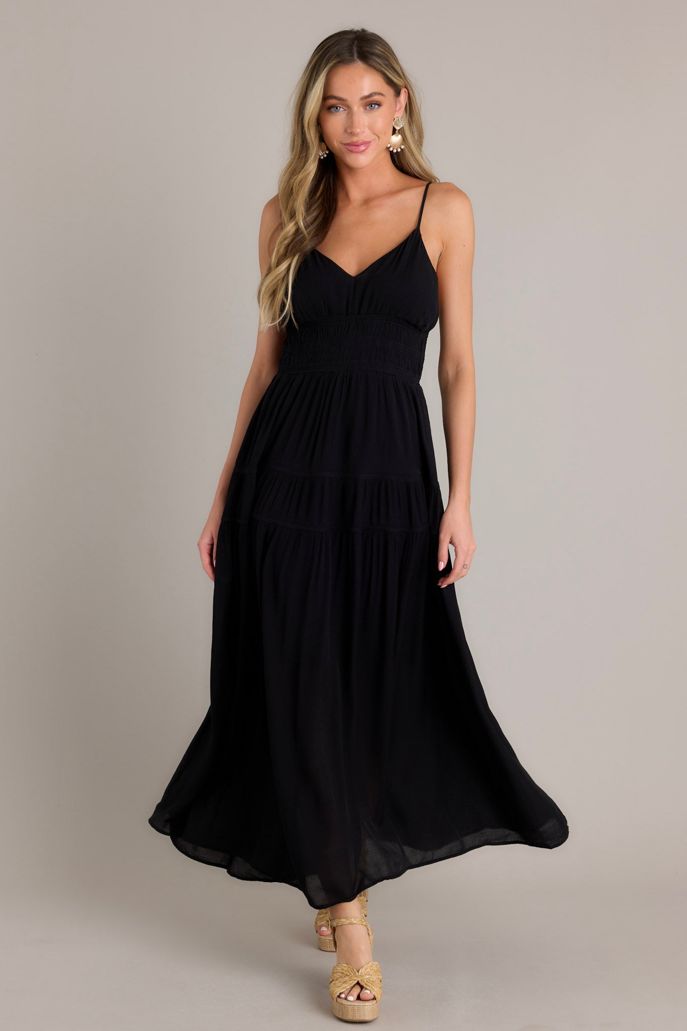 Full length view of a model wearing a black maxi dress with a v-neckline, thin adjustable straps, open back, fully smocked waist and lower back, tiers, and a flowing silhouette.