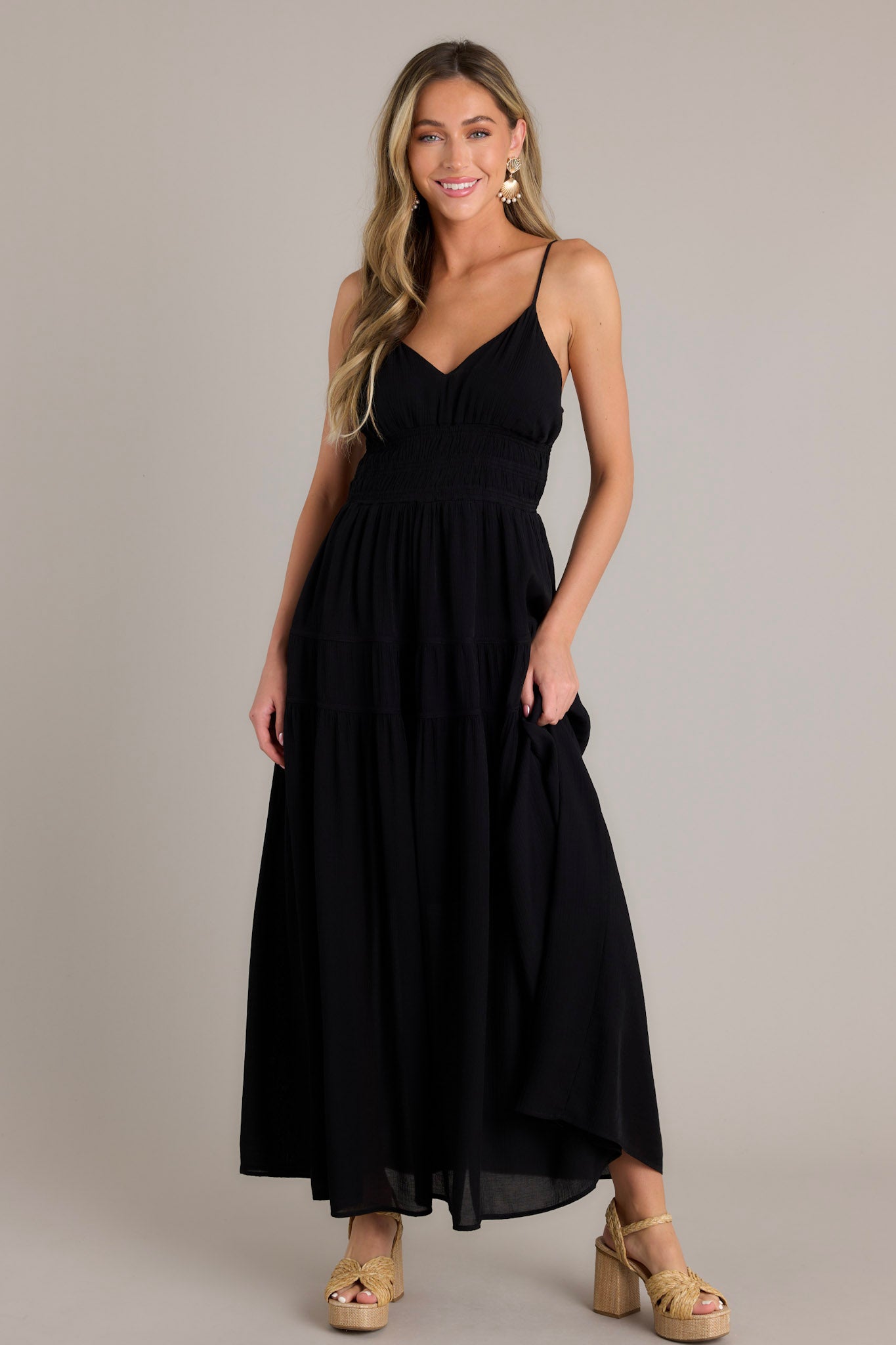 Action shot of a black maxi dress displaying the flow and movement of the fabric, highlighting the v-neckline, thin adjustable straps, open back, fully smocked waist and lower back, tiers, and a flowing silhouette.