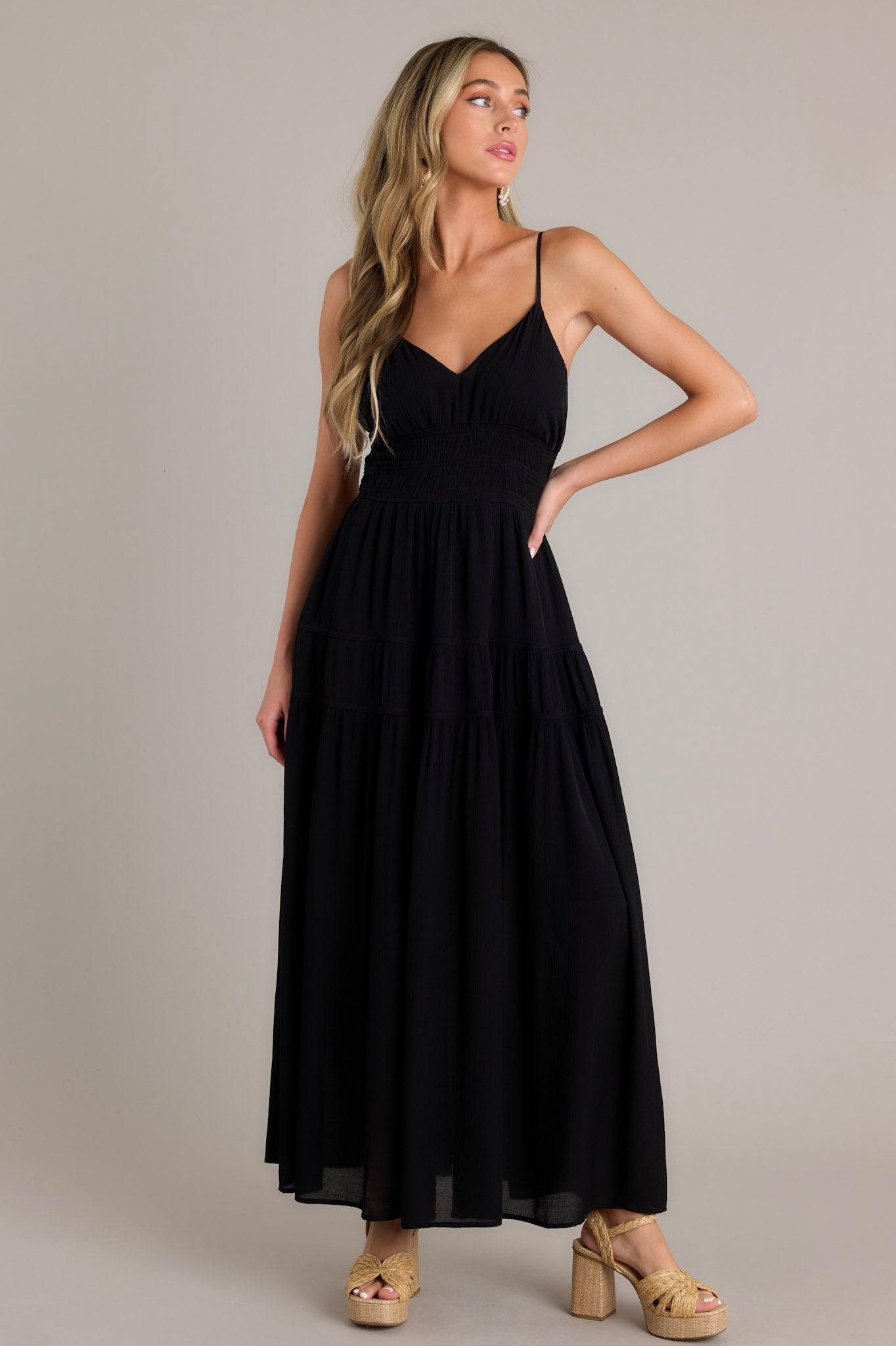 Front angled view of a black maxi dress featuring a v-neckline, thin adjustable straps, open back, fully smocked waist and lower back, tiers, and a flowing silhouette