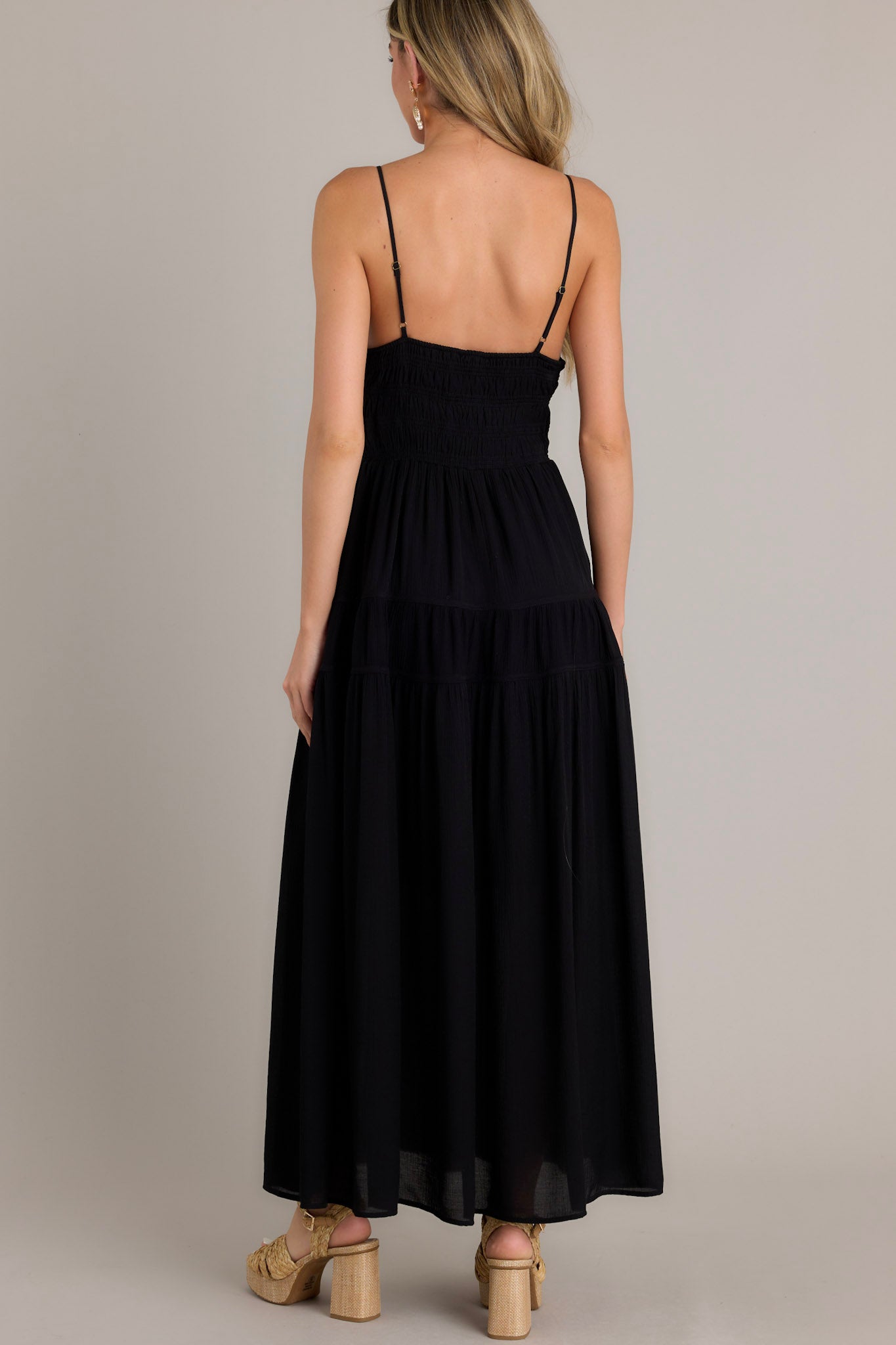 Back view of a black maxi dress highlighting the open back, thin adjustable straps, fully smocked waist and lower back, tiers, and a flowing silhouette.