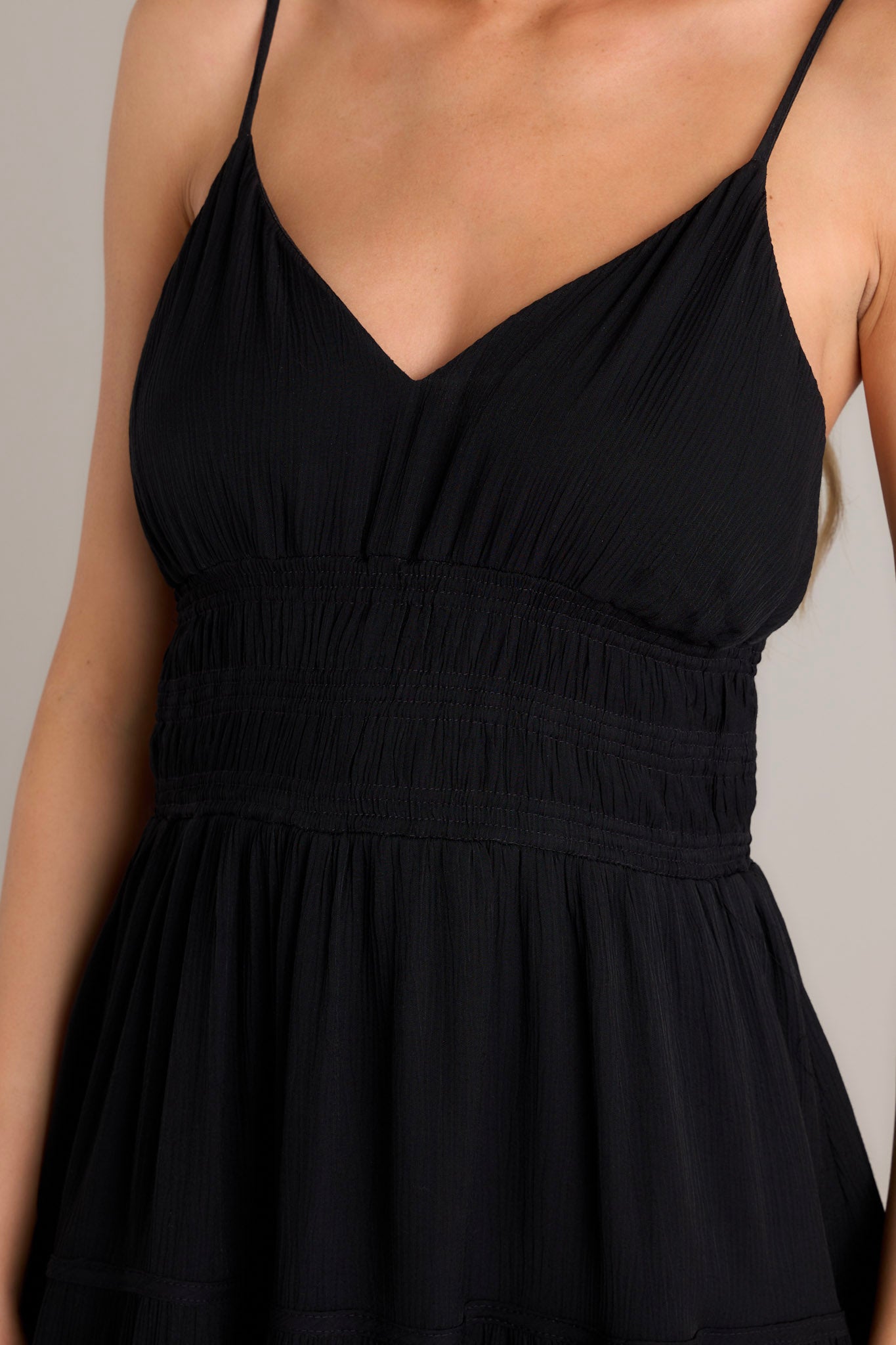Close-up of the black maxi dress showing the v-neckline, thin adjustable straps, smocked waist and lower back details, and tiered fabric.