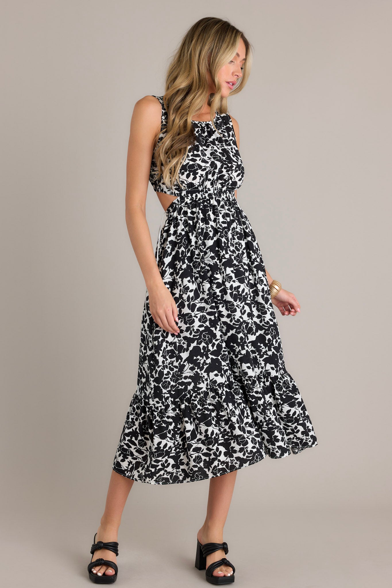 Front angled view of a black midi dress featuring a round neckline, self-tie feature at the back of the neck, an elastic waistband, waist cutouts, an open lower back, a single tier, and a flowing silhouette