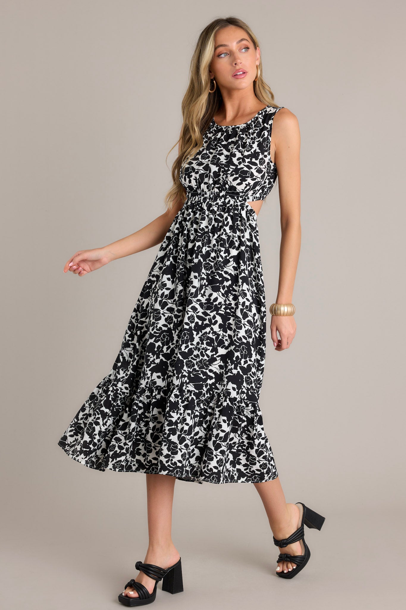 Action shot of a black midi dress displaying the flow and movement of the fabric, highlighting the round neckline, self-tie feature at the back of the neck, waist cutouts, an elastic waistband, an open lower back, a single tier, and a flowing silhouette.