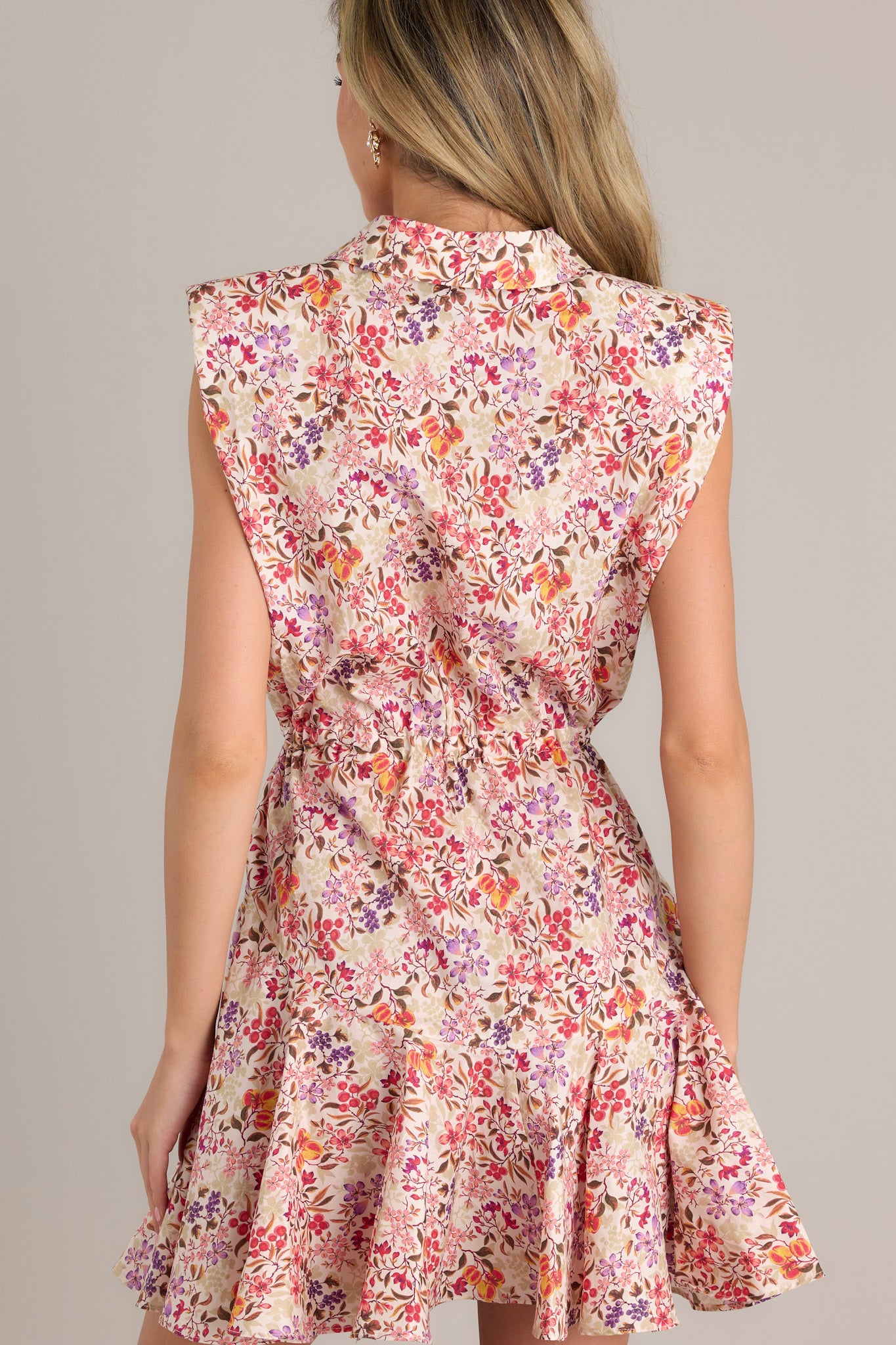 Back view of the beige floral multi mini dress featuring sleeveless padded shoulders, a collared neckline, and a flowy ruffled skirt with an elastic waist accentuated with tassels.