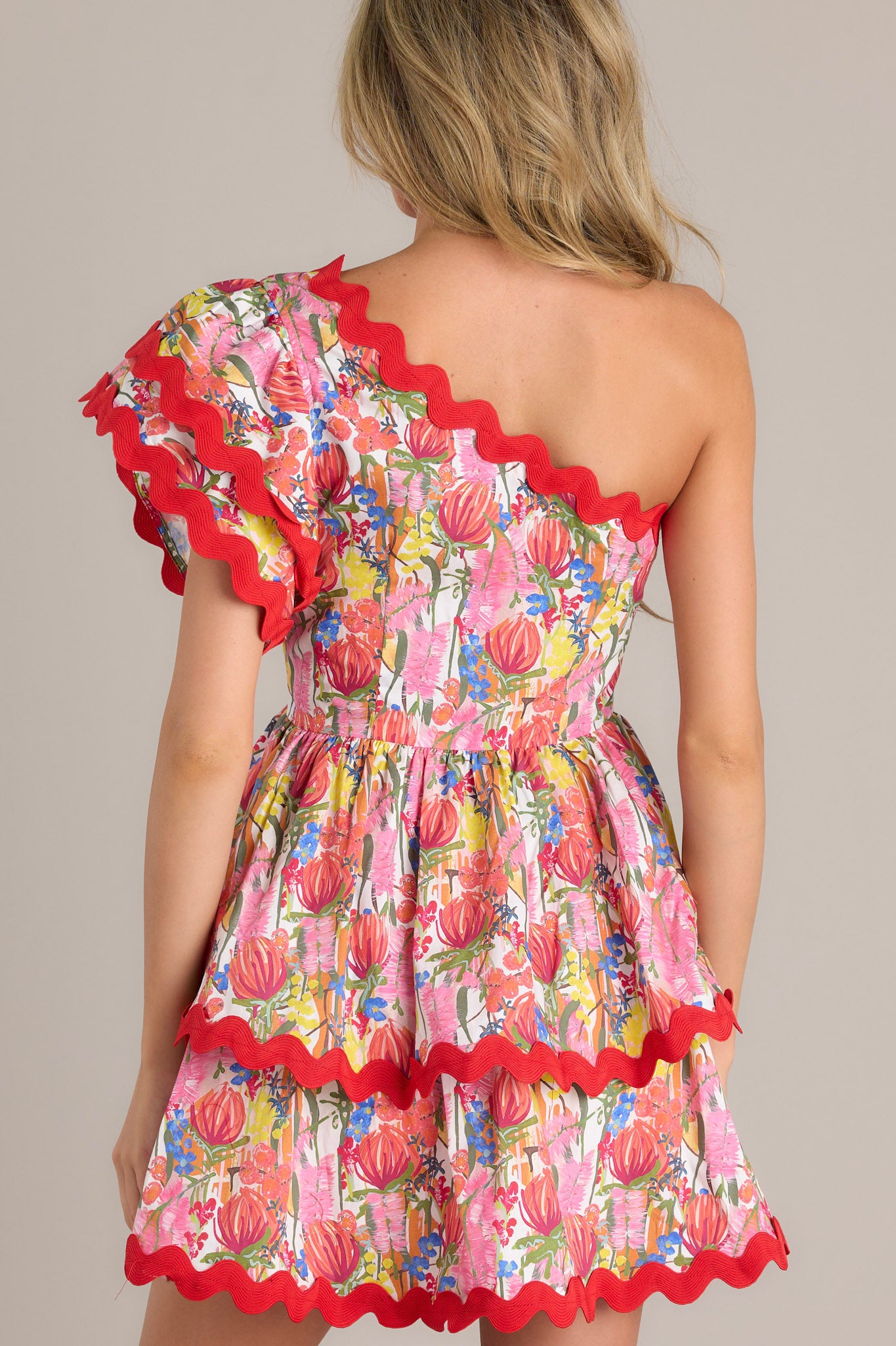 Back view of this red multi floral romper, featuring a one shoulder neckline, fitted bodice with elastic waist, and tiered skirt.