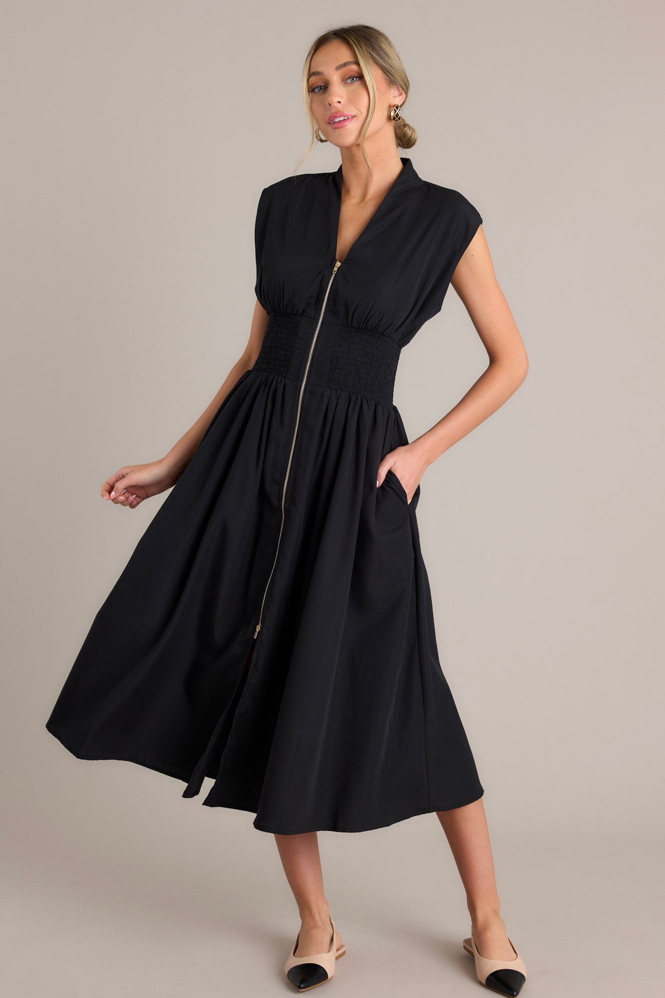 Action shot of a black midi dress displaying the fit and movement, highlighting the v-neckline, padded shoulders, full zipper front, smocked waist, functional hip pockets, and front slit.