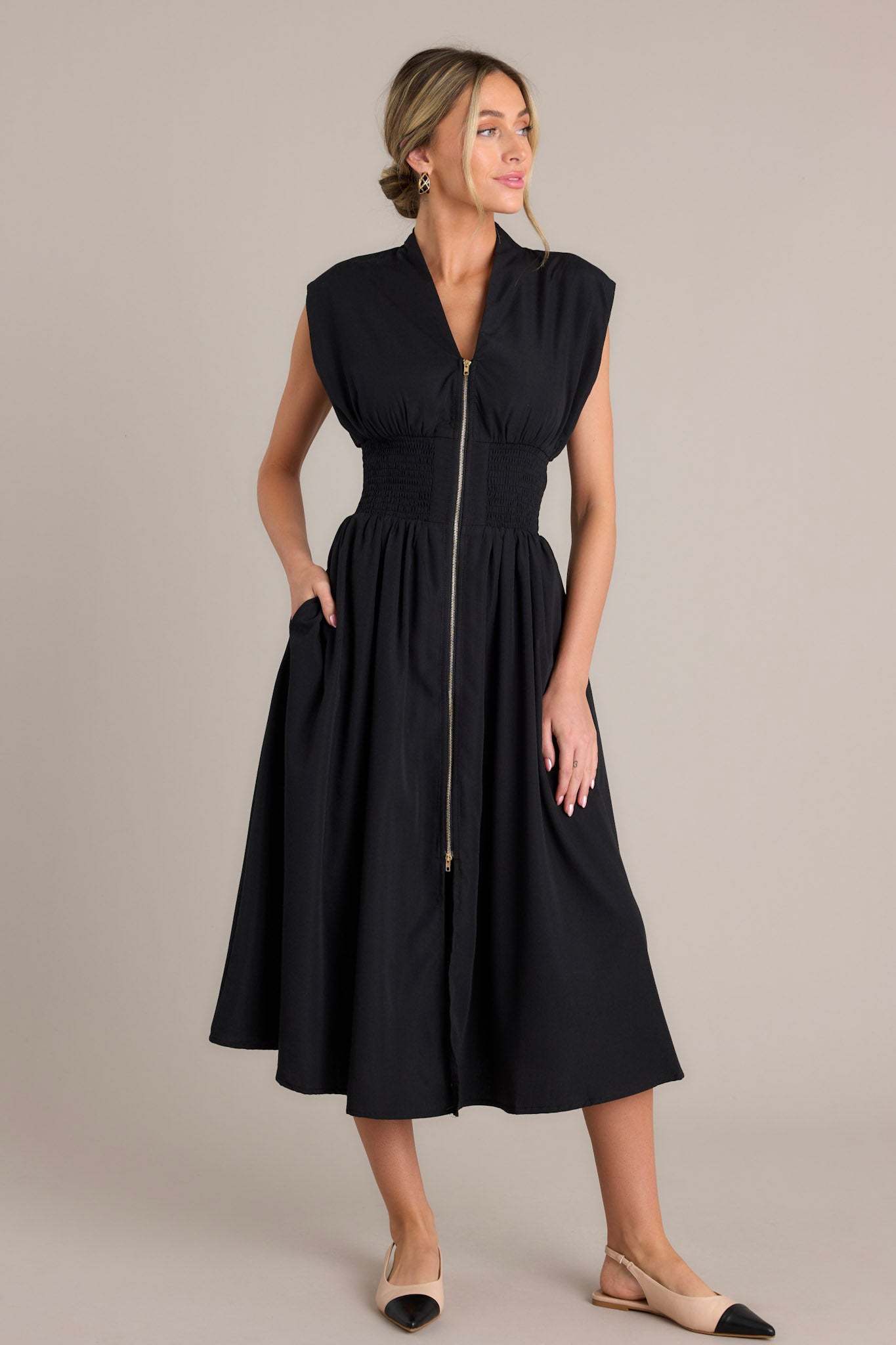 Full length view of a black midi dress with a v-neckline, padded shoulders, a full zipper front, a fully smocked waist, functional hip pockets, and a front slit