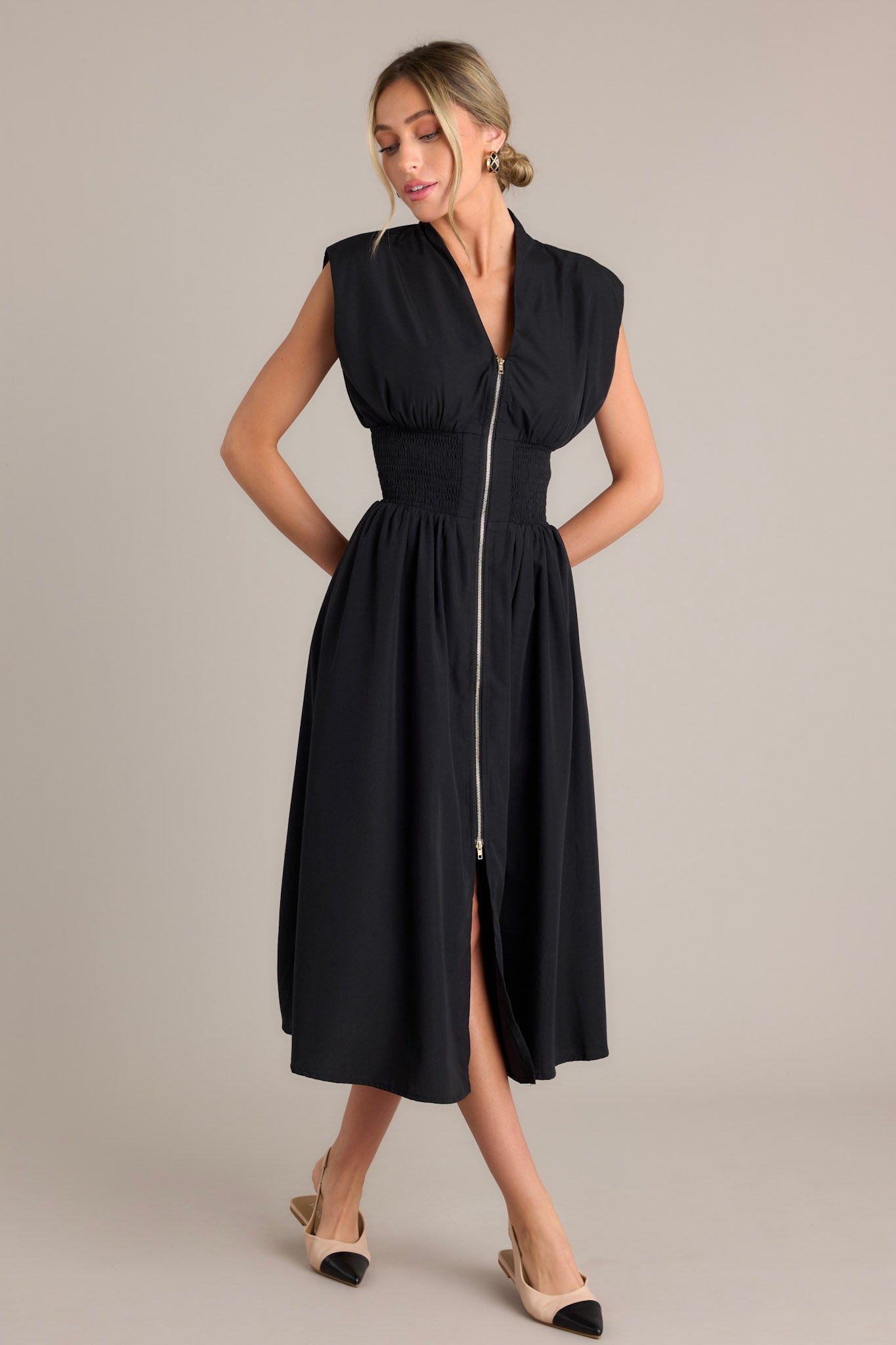 Front angled view of a black midi dress featuring a v-neckline, padded shoulders, a full zipper front, a fully smocked waist, functional hip pockets, and a front slit
