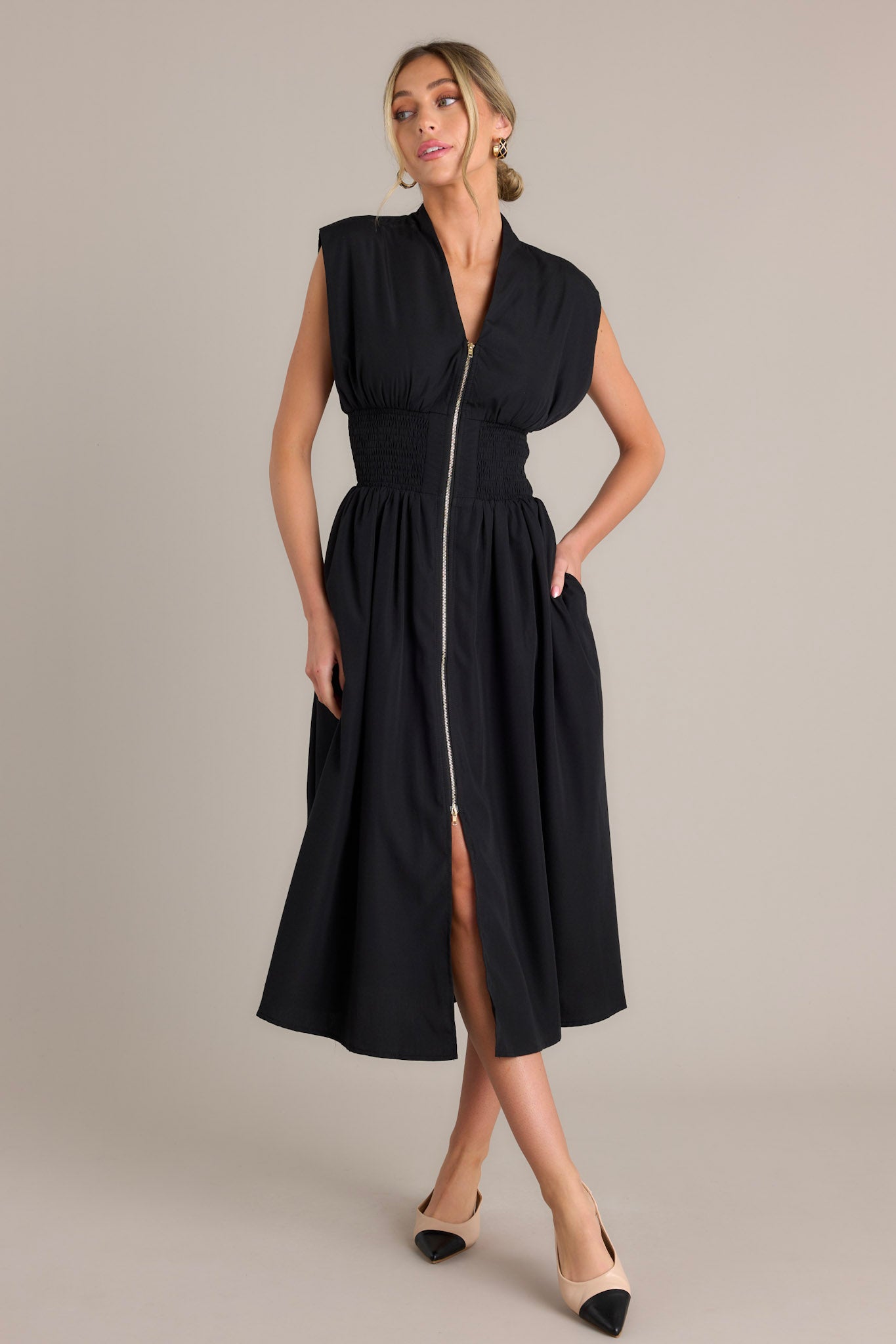 This black midi dress features a v-neckline, padded shoulders, a full zipper front, a fully smocked waist, functional hip pockets, and a front slit.